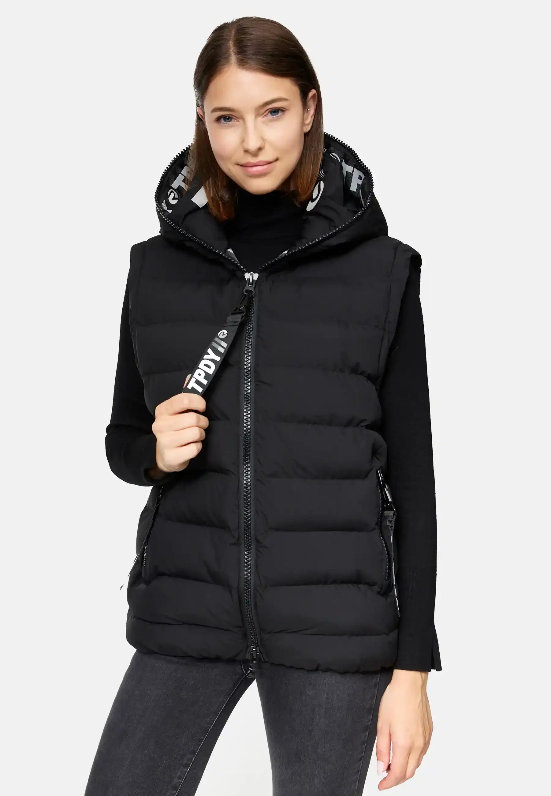 Eine Frau mit glattem braunem Haar trägt die trueprodigy Matthew F Damenweste mit Kapuze matt- eine schwarze Kapuzenweste- über einem schwarzen langärmeligen Oberteil und dunklen Jeans. Sie hält den Reißverschluss zu und blickt in die Kamera vor einem schlichten Hintergrund.