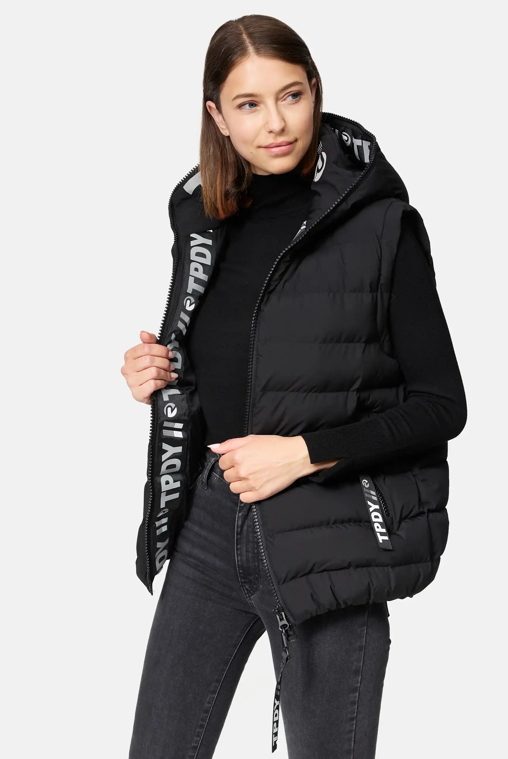 Eine Frau trägt eine trueprodigy Matthew F Damenweste mit Kapuze matt über einem schwarzen langärmeligen Oberteil und einer dunklen Jeans. Sie steht vor einem schlichten Hintergrund und schaut leicht nach links.