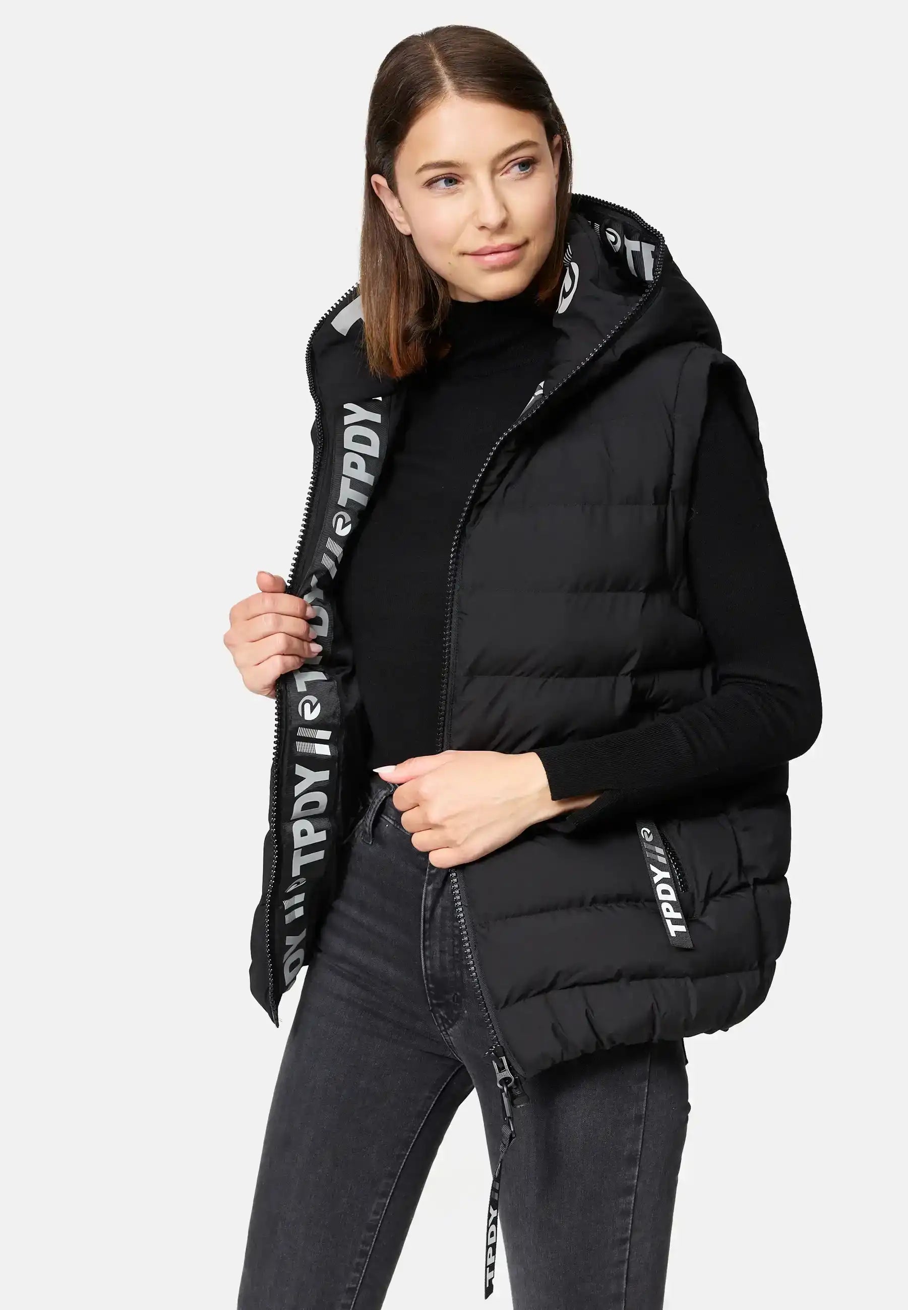 Eine Frau trägt eine trueprodigy Matthew F Damenweste mit Kapuze matt über einem schwarzen langärmeligen Oberteil und einer dunklen Jeans. Sie steht vor einem schlichten Hintergrund und schaut leicht nach links.
