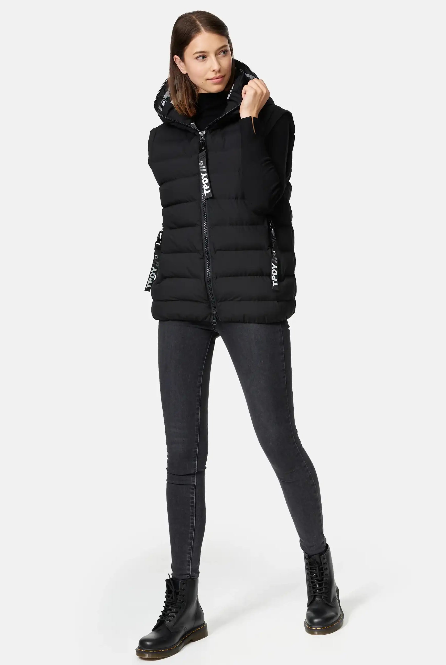 Eine Frau steht vor einem schlichten Hintergrund und trägt die Matthew F Damenweste mit Kapuze matt von trueprodigy über einem schwarzen langärmeligen Oberteil, dunklen Skinny Jeans und schwarzen Schnürstiefeln. Sie hält den Kragen der Weste mit einer Hand und blickt leicht zur Seite.