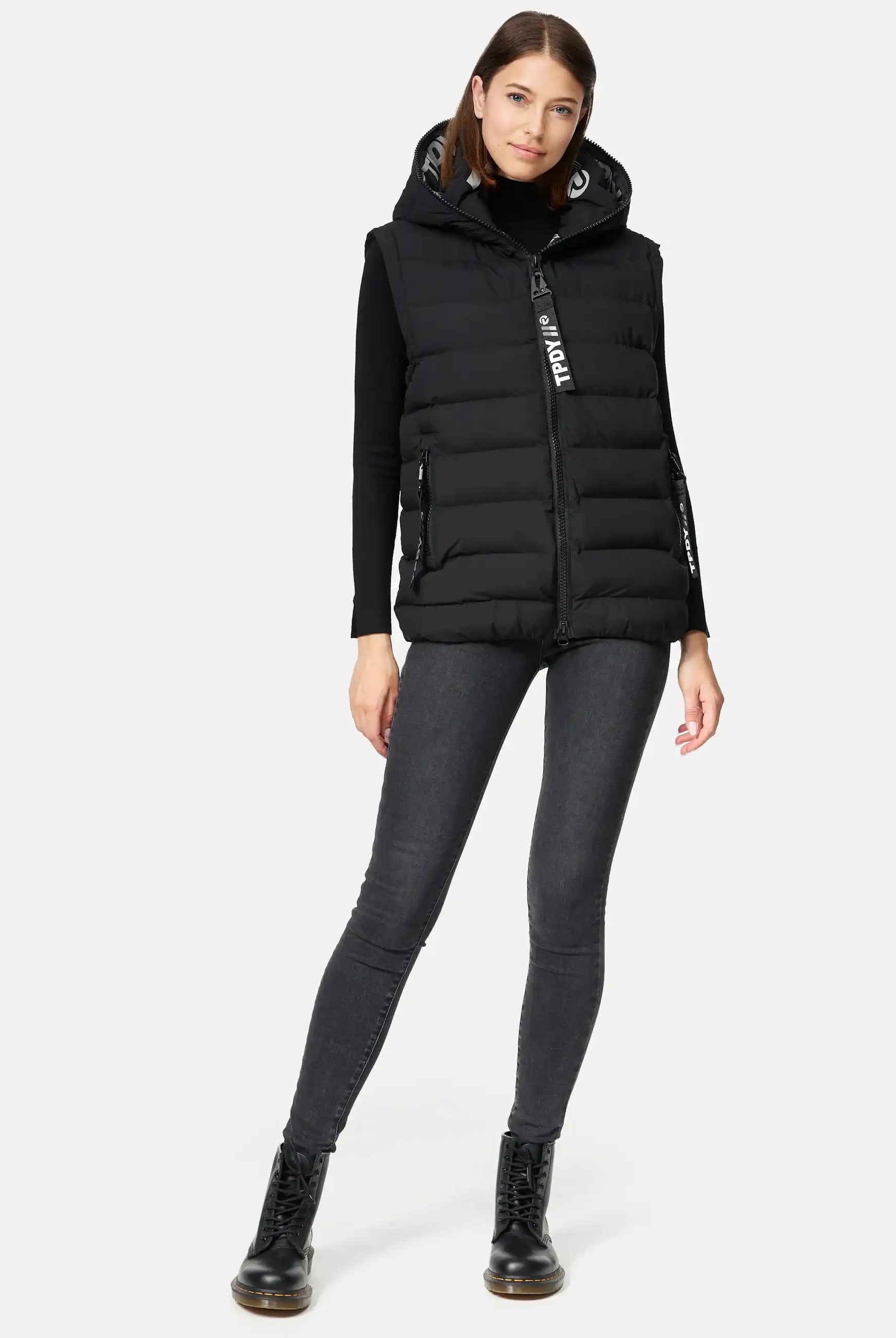 Eine Frau steht vor einem schlichten weißen Hintergrund und trägt eine trueprodigy Matthew F Damenweste mit Kapuze matt, ein schwarzes langärmeliges Oberteil, dunkle Skinny Jeans und schwarze Schnürstiefel.