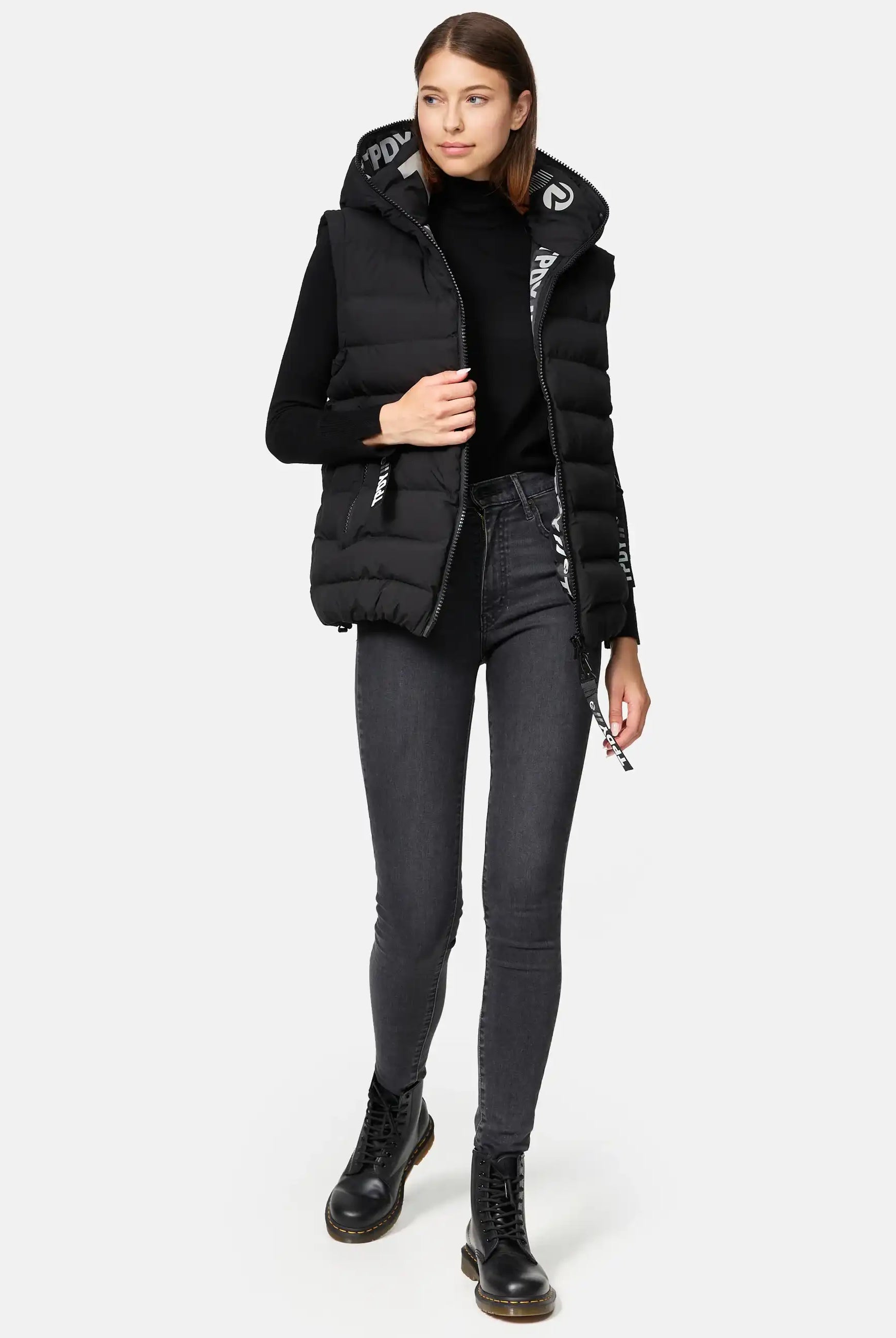 Eine Frau mit glattem braunem Haar und neutralem Gesichtsausdruck steht vor einem schlichten Hintergrund und trägt die trueprodigy Matthew F Damenweste mit Kapuze matt, einen schwarzen Rollkragenpullover, dunkelgraue Jeans und schwarze Schnürstiefel.