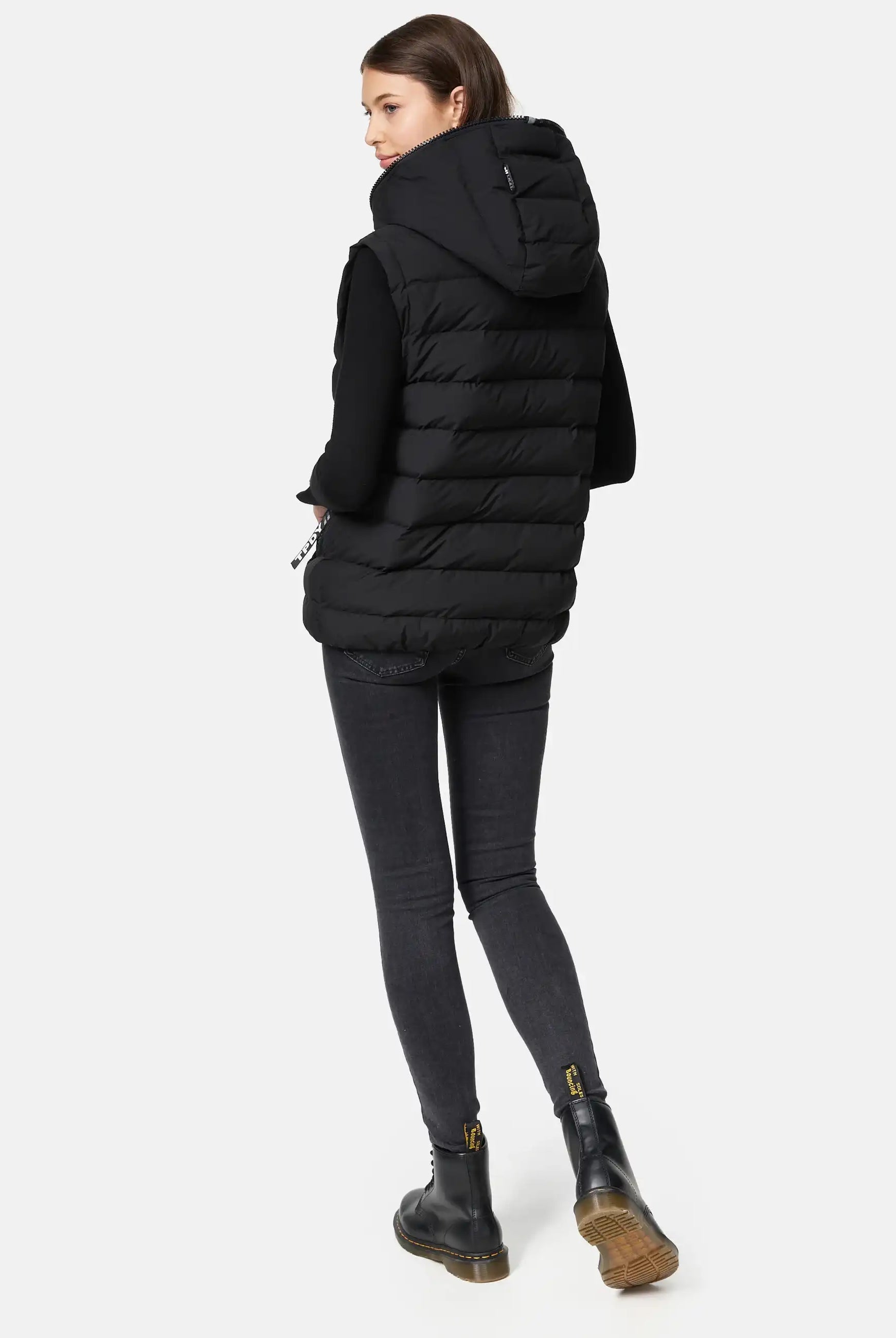 Eine Person steht mit dem Gesicht leicht abgewandt und trägt ein mattes, schwarzes, langärmeliges Oberteil, dunkle Skinny-Jeans und schwarze Ankle Boots mit gelben Laschen vor einem hellgrauen Hintergrund.