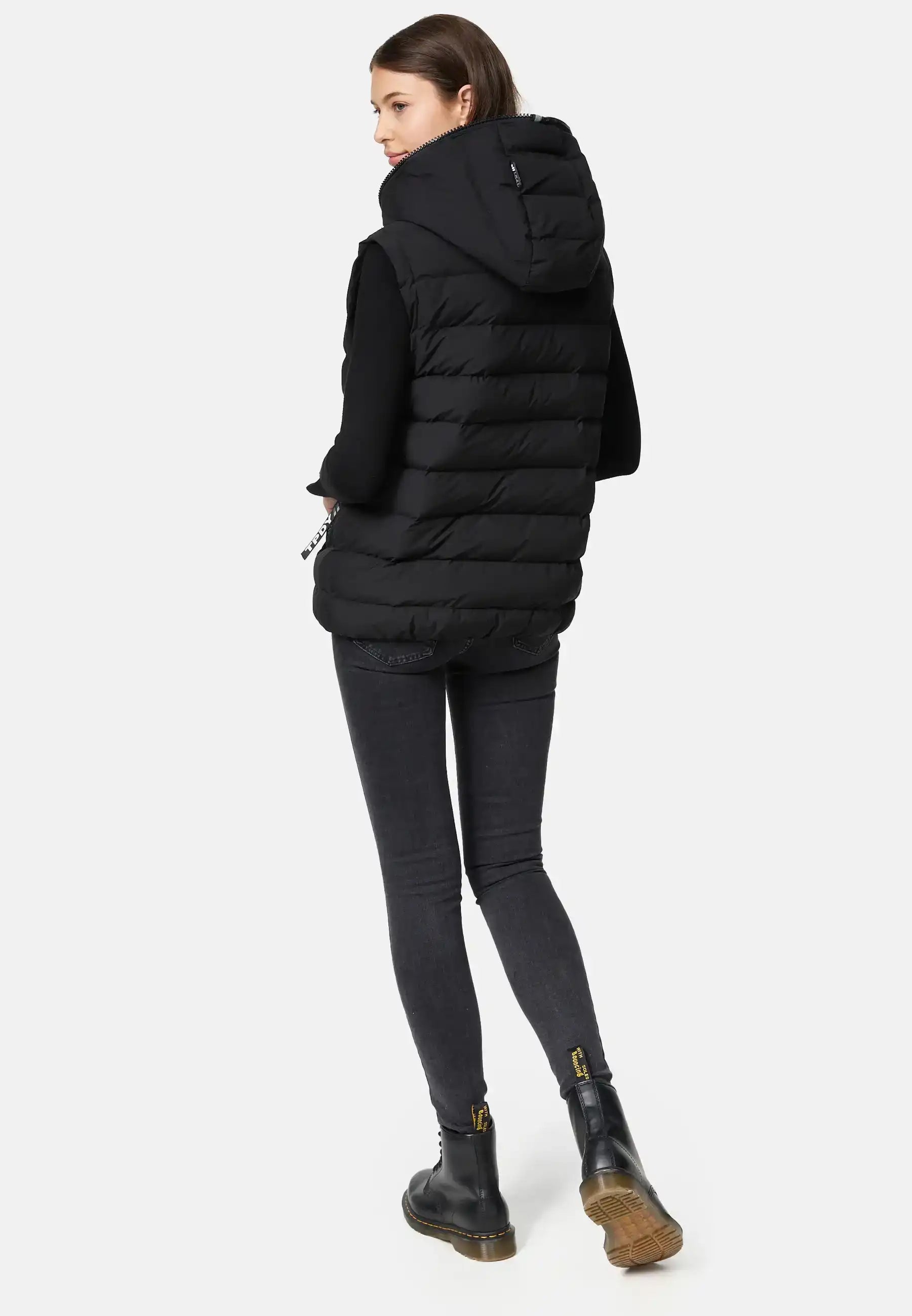 Eine Person steht mit dem Gesicht leicht abgewandt und trägt ein mattes, schwarzes, langärmeliges Oberteil, dunkle Skinny-Jeans und schwarze Ankle Boots mit gelben Laschen vor einem hellgrauen Hintergrund.