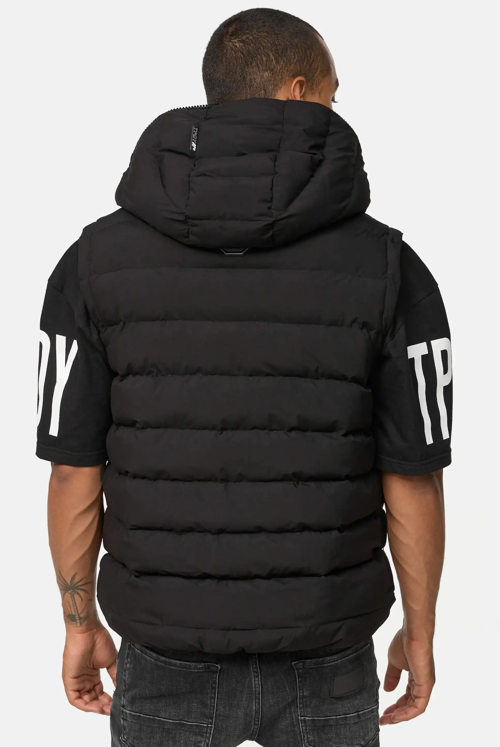 Eine Person mit kurzen Haaren und einer Tätowierung auf dem linken Unterarm wendet sich von der Kamera ab. Sie trägt eine schwarze Trueprodigy Matthew Pufferweste mit Kapuze matt über einem schwarzen kurzärmeligen Hemd mit weißem Ärmelaufdruck und einer verblichenen schwarzen Jeans.