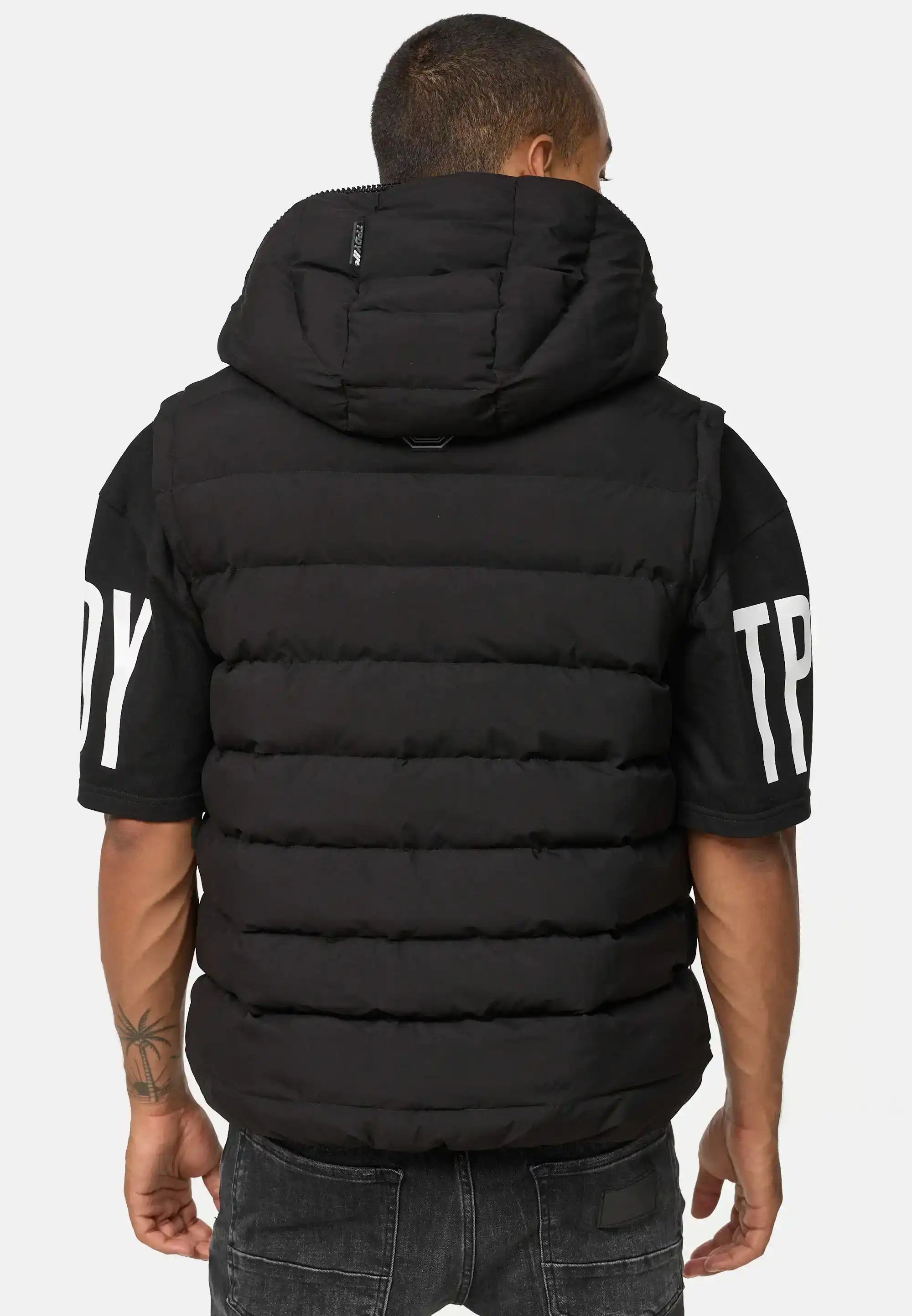 Eine Person mit kurzen Haaren und einer Tätowierung auf dem linken Unterarm wendet sich von der Kamera ab. Sie trägt eine schwarze Trueprodigy Matthew Pufferweste mit Kapuze matt über einem schwarzen kurzärmeligen Hemd mit weißem Ärmelaufdruck und einer verblichenen schwarzen Jeans.