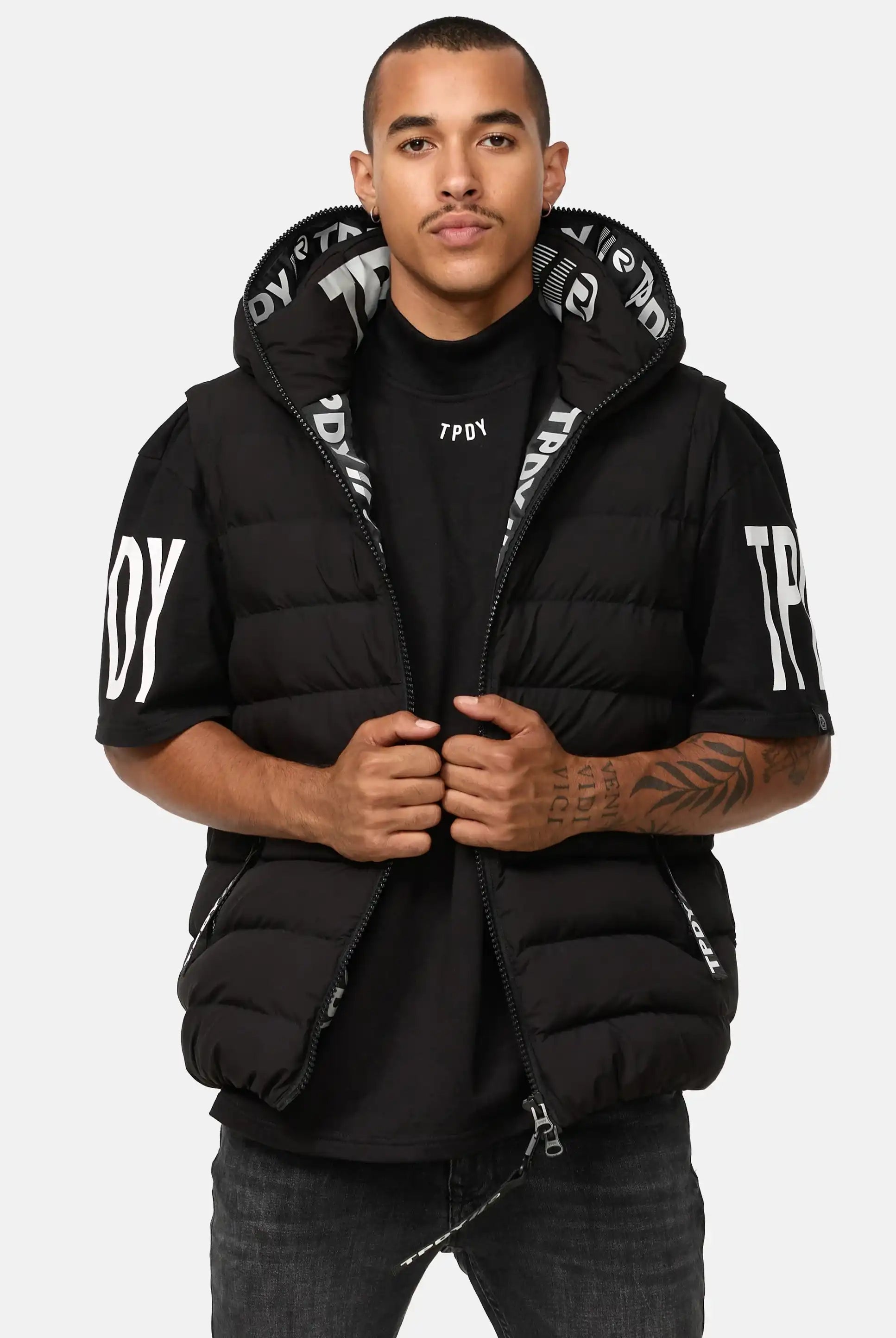 Ein Mann mit kurzen Haaren und einer Unterarmtätowierung steht vor der Kamera. Er trägt eine schwarze Matthew Pufferweste mit Kapuze matt über einem T-Shirt mit weißem Schriftzug und hält die Weste mit geöffnetem Reißverschluss offen, so dass der innere Text zu sehen ist, während die Kapuze geöffnet ist.