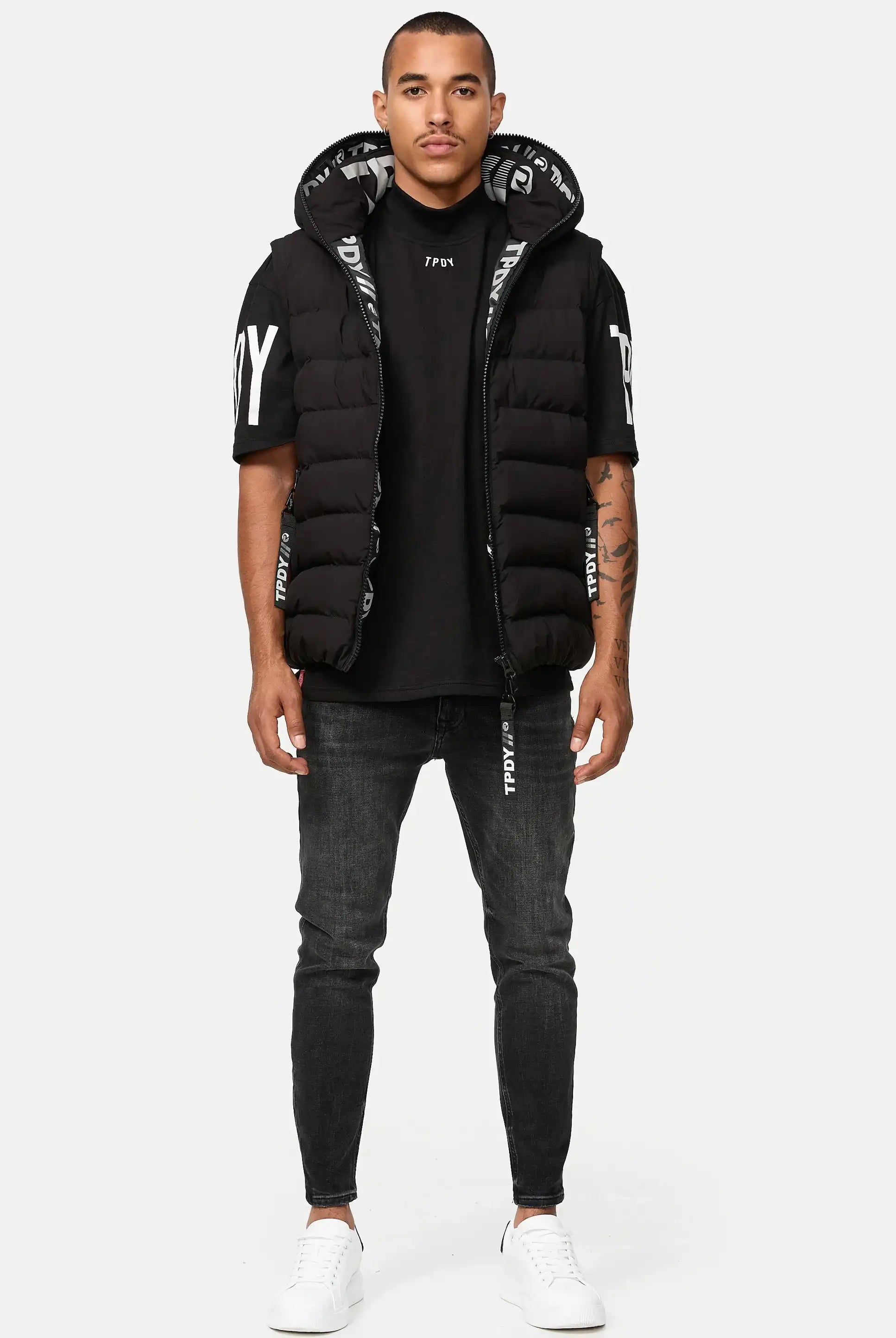 Ein Mann steht mit dem Gesicht nach vorne in einer Trueprodigy Matthew Pufferweste mit Kapuze matt, einem schwarzen T-Shirt mit weißem Text, dunklen Skinny Jeans und weißen Turnschuhen vor einem schlichten weißen Hintergrund.