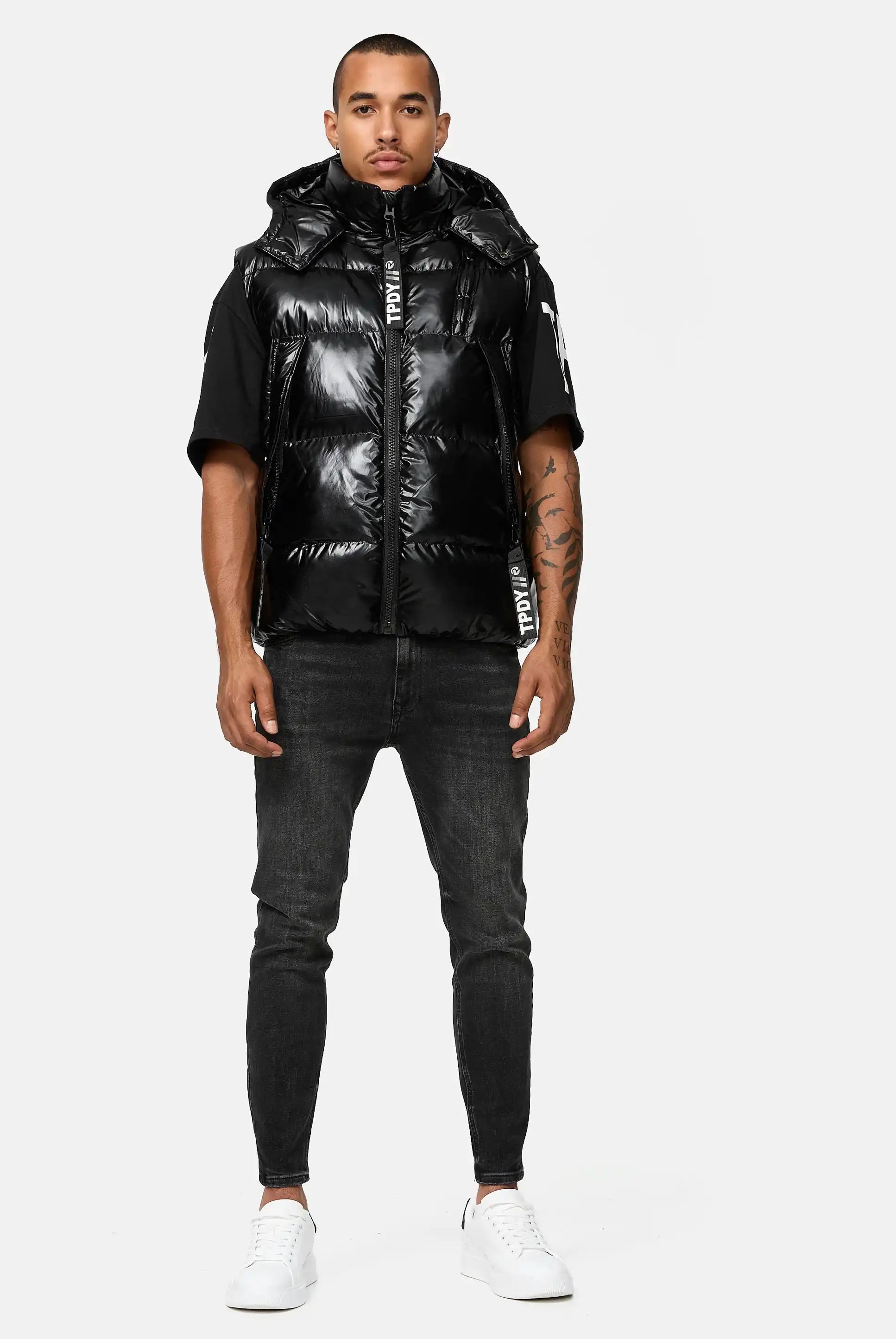 Ein Mann blickt nach vorne und trägt die Trueprodigy Darko Steppweste glänzend mit Kapuze über einem schwarzen kurzärmeligen Hemd, dunkle Skinny Jeans und weiße Turnschuhe vor einem schlichten, hellen Hintergrund.