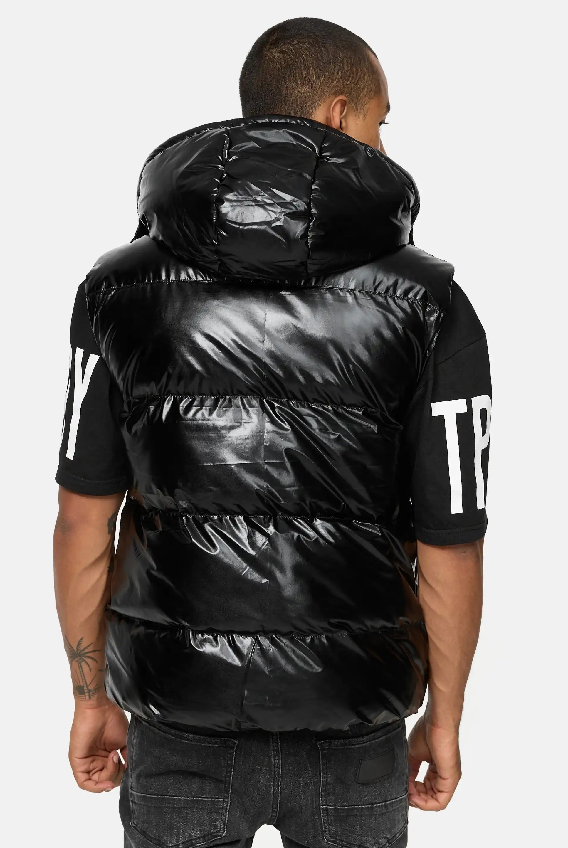 Von hinten ist eine Person zu sehen, die eine schwarze Darko Steppweste glänzend mit Kapuze von trueprodigy über einem schwarzen T-Shirt mit weißem Schriftzug an den Ärmeln trägt, gepaart mit einer dunklen Jeans.