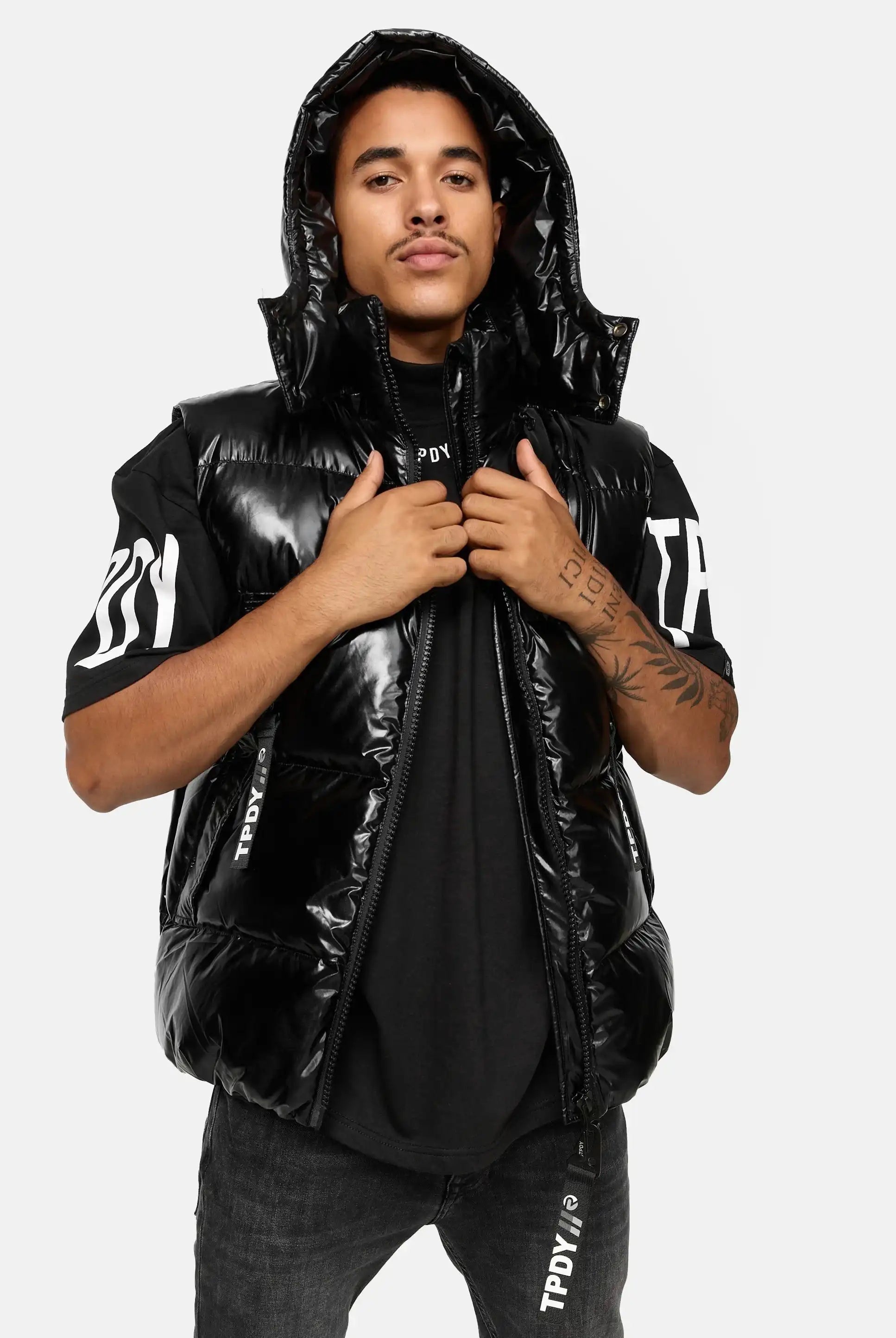 Eine Person trägt die glänzende Darko Steppweste mit Kapuze von trueprodigy über einem schwarzen T-Shirt mit weißem Text auf Ärmeln und Brust sowie dunkler Jeans und steht dabei vor hellem Hintergrund.