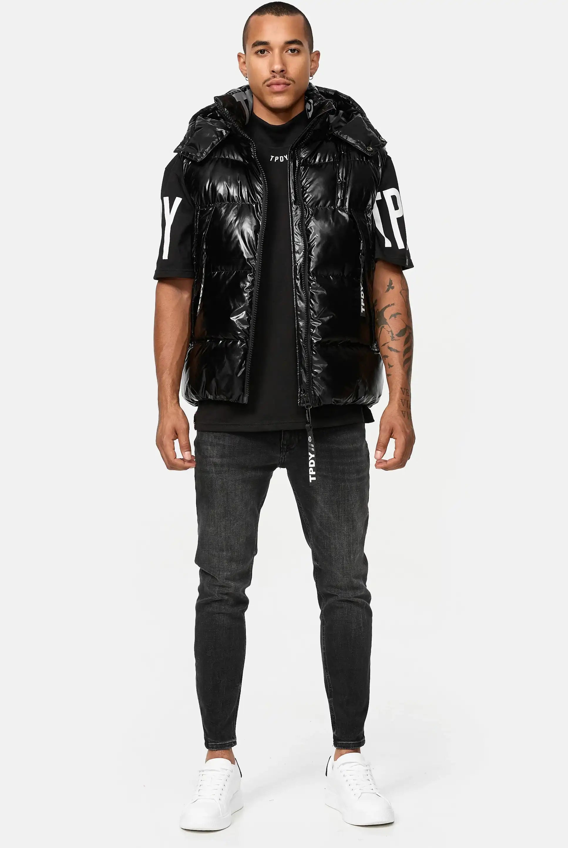 Ein Mann blickt nach vorne und trägt die Trueprodigy Darko Steppweste glänzend mit Kapuze über einem schwarzen T-Shirt mit weißem Ärmelaufdruck, dunkle Skinny Jeans und weiße Turnschuhe vor einem schlichten weißen Hintergrund.