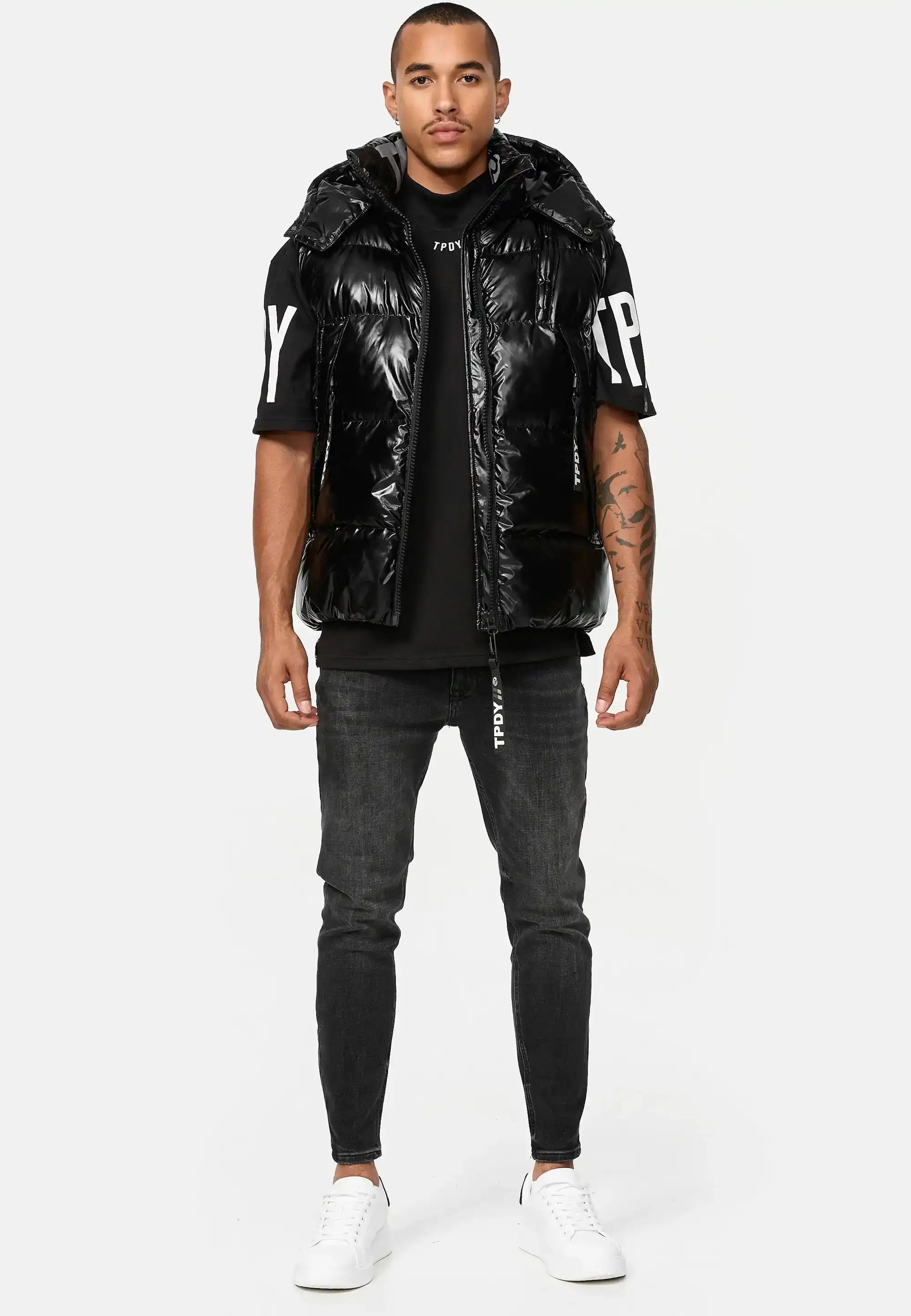 Ein Mann blickt nach vorne und trägt die Trueprodigy Darko Steppweste glänzend mit Kapuze über einem schwarzen T-Shirt mit weißem Ärmelaufdruck, dunkle Skinny Jeans und weiße Turnschuhe vor einem schlichten weißen Hintergrund.