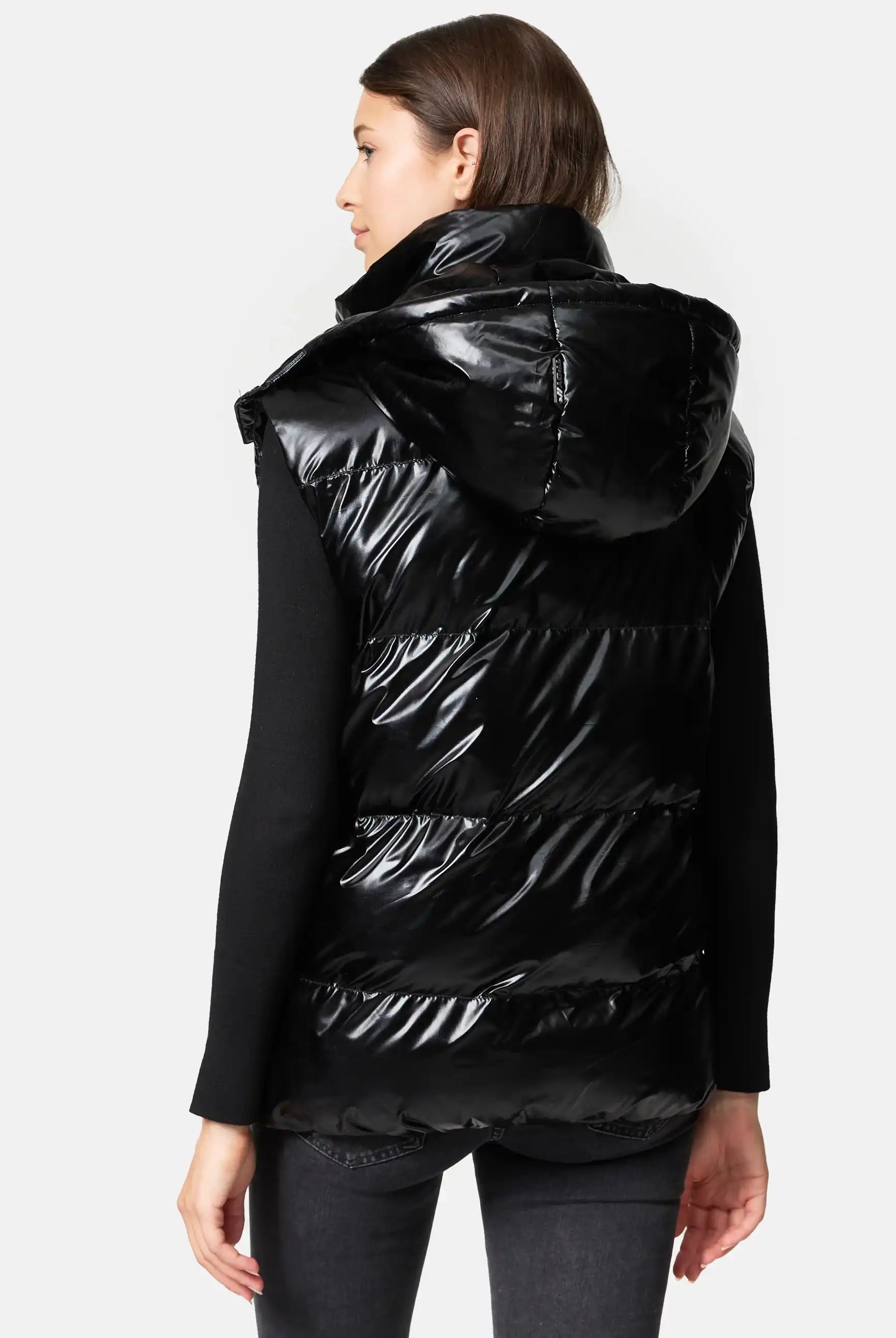 Eine Person steht mit dem Gesicht nach unten und trägt die Trueprodigy Darko F Weste glänzend mit Kapuze über einem schwarzen langärmeligen Oberteil und einer dunklen Hose. Der Hintergrund ist einfarbig und hell gefärbt.