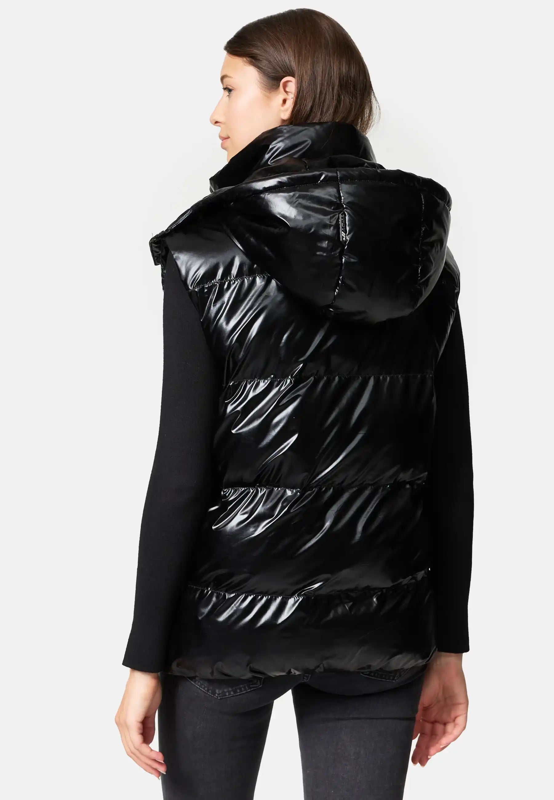 Eine Person steht mit dem Gesicht nach unten und trägt die Trueprodigy Darko F Weste glänzend mit Kapuze über einem schwarzen langärmeligen Oberteil und einer dunklen Hose. Der Hintergrund ist einfarbig und hell gefärbt.