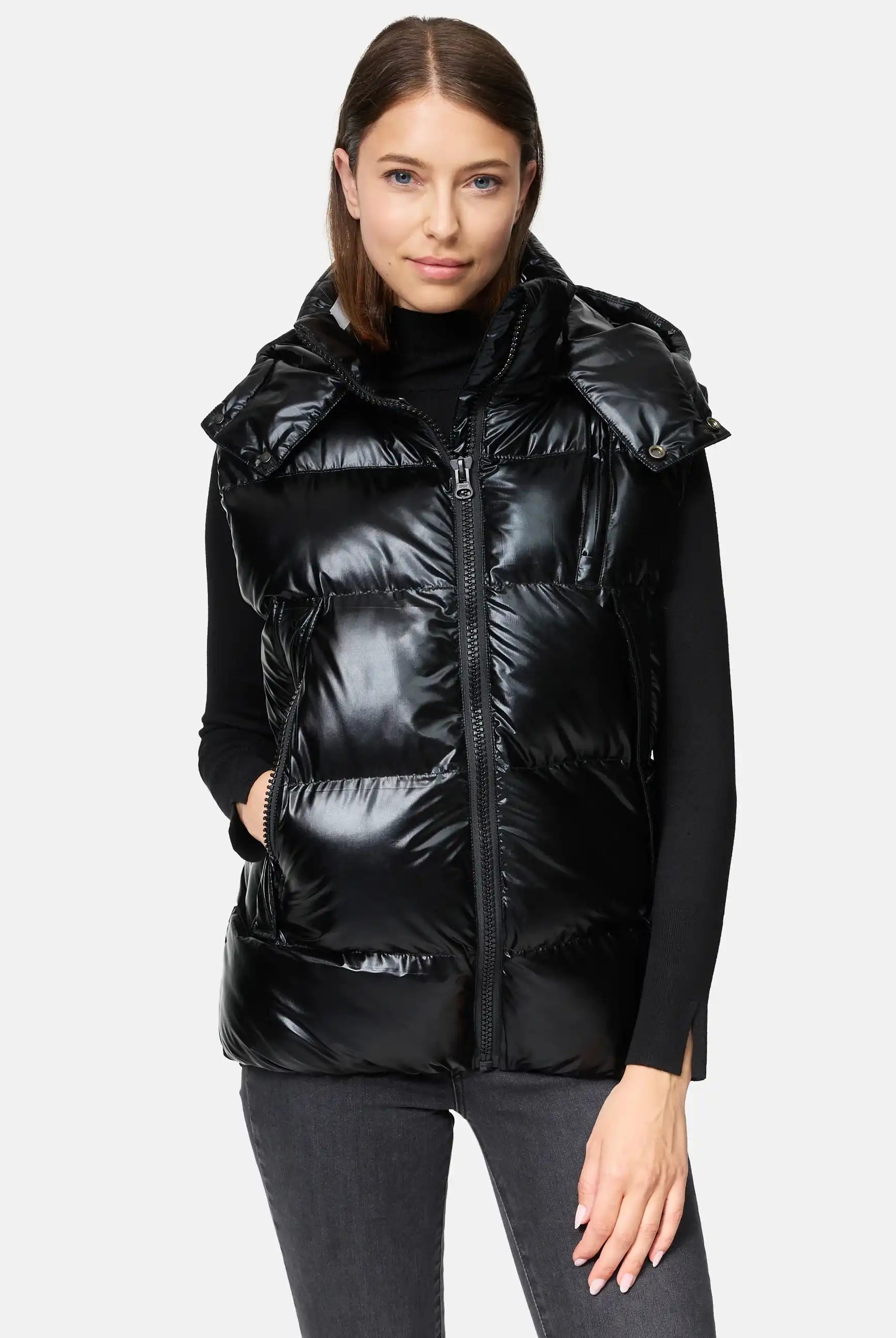 Eine Frau mit glattem braunem Haar trägt die trueprodigy Darko F Weste glänzend mit Kapuze über einem schwarzen Rollkragenpullover und grauen Jeans, eine Hand in der Westentasche, vor einem schlichten hellen Hintergrund.