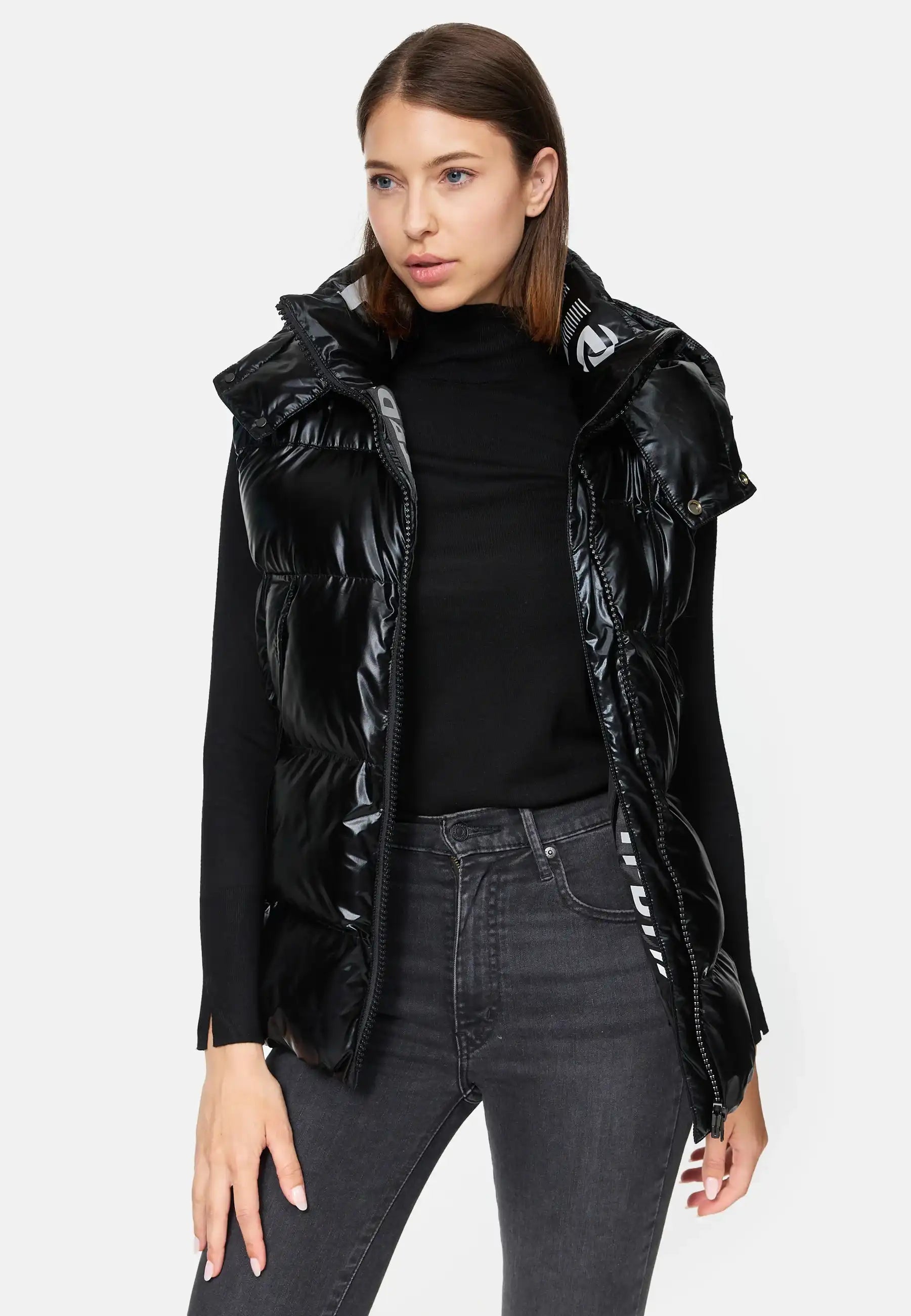 Eine Frau trägt die glänzende Darko F Weste mit Kapuze von trueprodigy offen über einem schwarzen Longsleeve-Turtleneck und dunklen Jeans. Ihr glattes Haar ist hinter die Ohren gesteckt, der Hintergrund ist schlicht weiß.