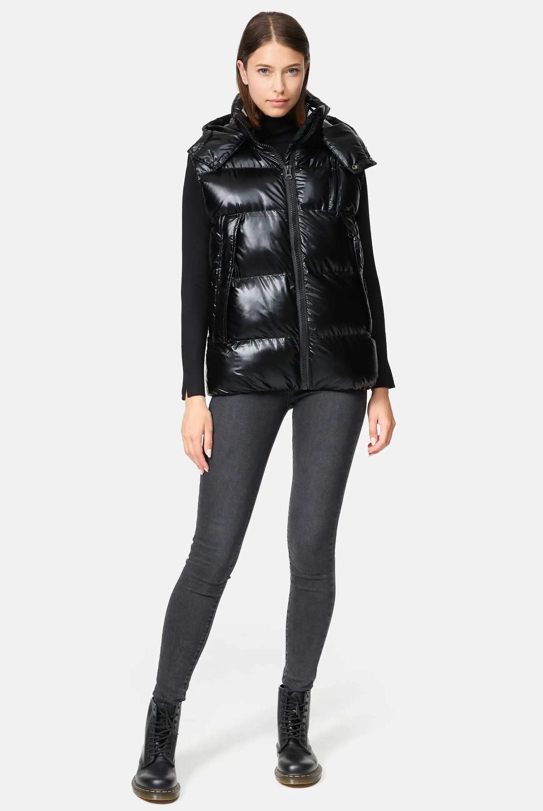 Eine Frau steht mit dem Gesicht nach vorne und trägt die Trueprodigy Darko F Weste glänzend mit Kapuze über einem schwarzen langärmeligen Oberteil, dunkelgrauen Skinny Jeans und schwarzen Schnürstiefeln vor einem schlichten hellen Hintergrund.