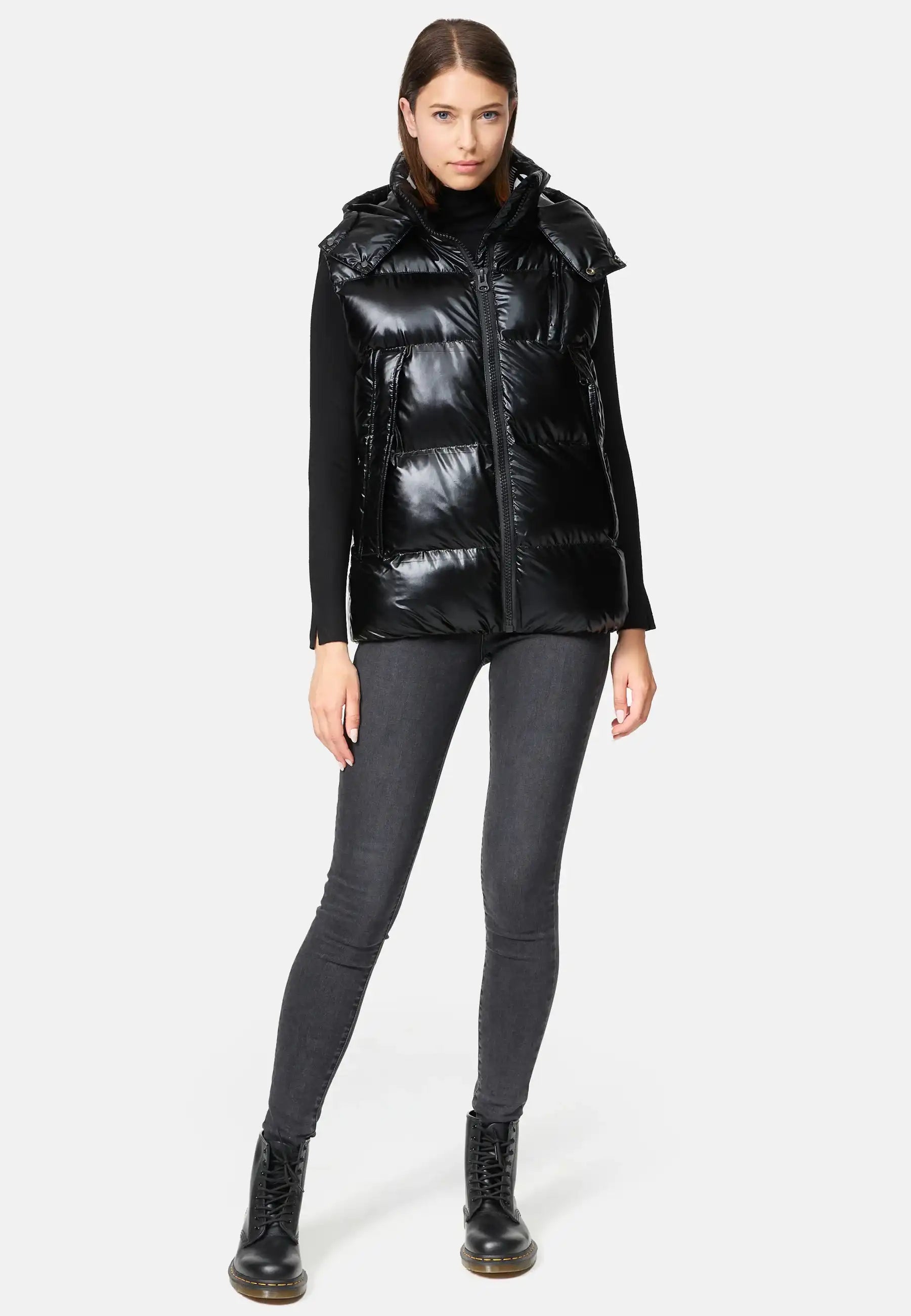 Eine Frau steht mit dem Gesicht nach vorne und trägt die Trueprodigy Darko F Weste glänzend mit Kapuze über einem schwarzen langärmeligen Oberteil, dunkelgrauen Skinny Jeans und schwarzen Schnürstiefeln vor einem schlichten hellen Hintergrund.