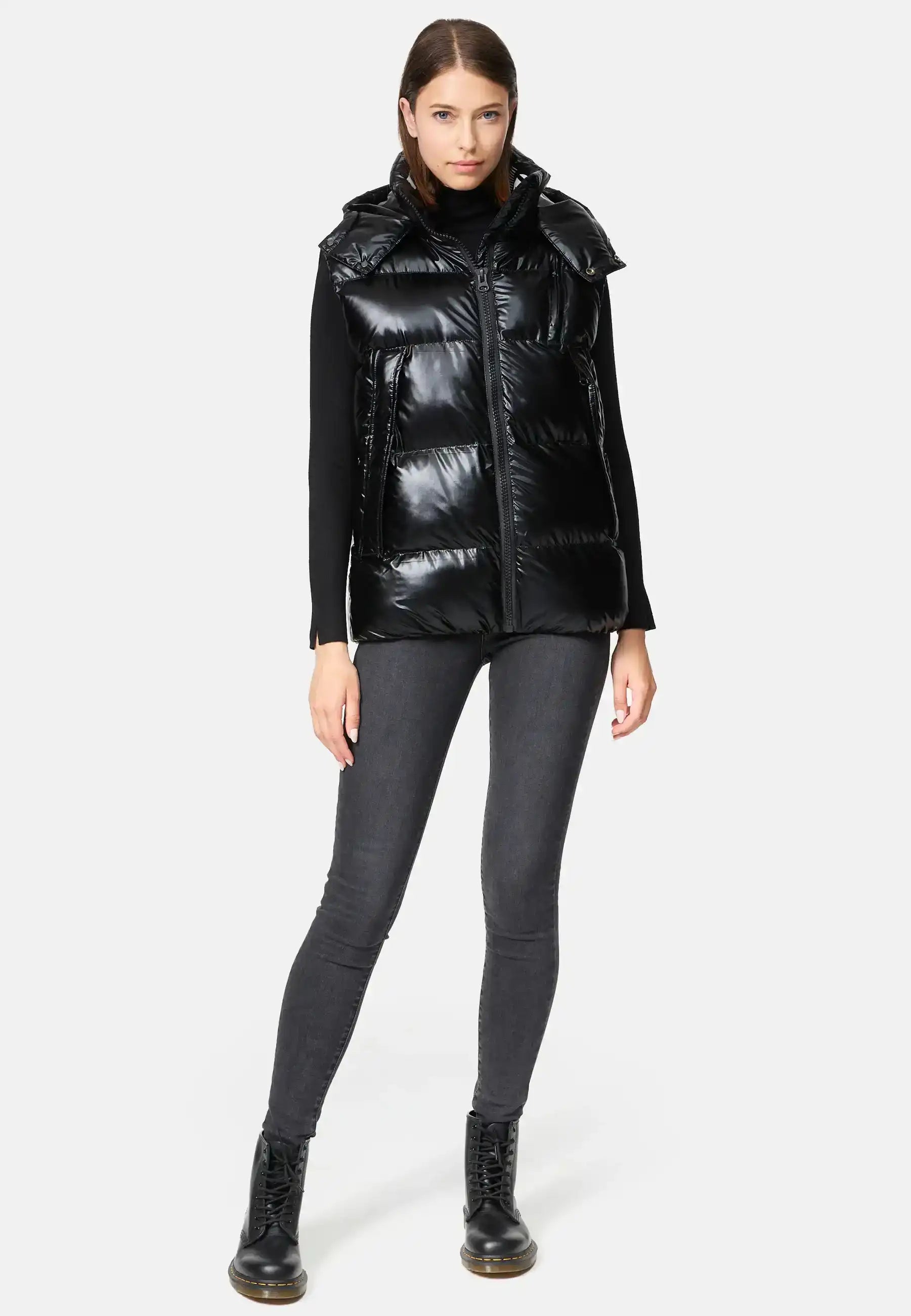Eine Frau steht mit dem Gesicht nach vorne und trägt die Trueprodigy Darko F Weste glänzend mit Kapuze über einem schwarzen langärmeligen Oberteil, dunkelgrauen Skinny Jeans und schwarzen Schnürstiefeln vor einem schlichten hellen Hintergrund.