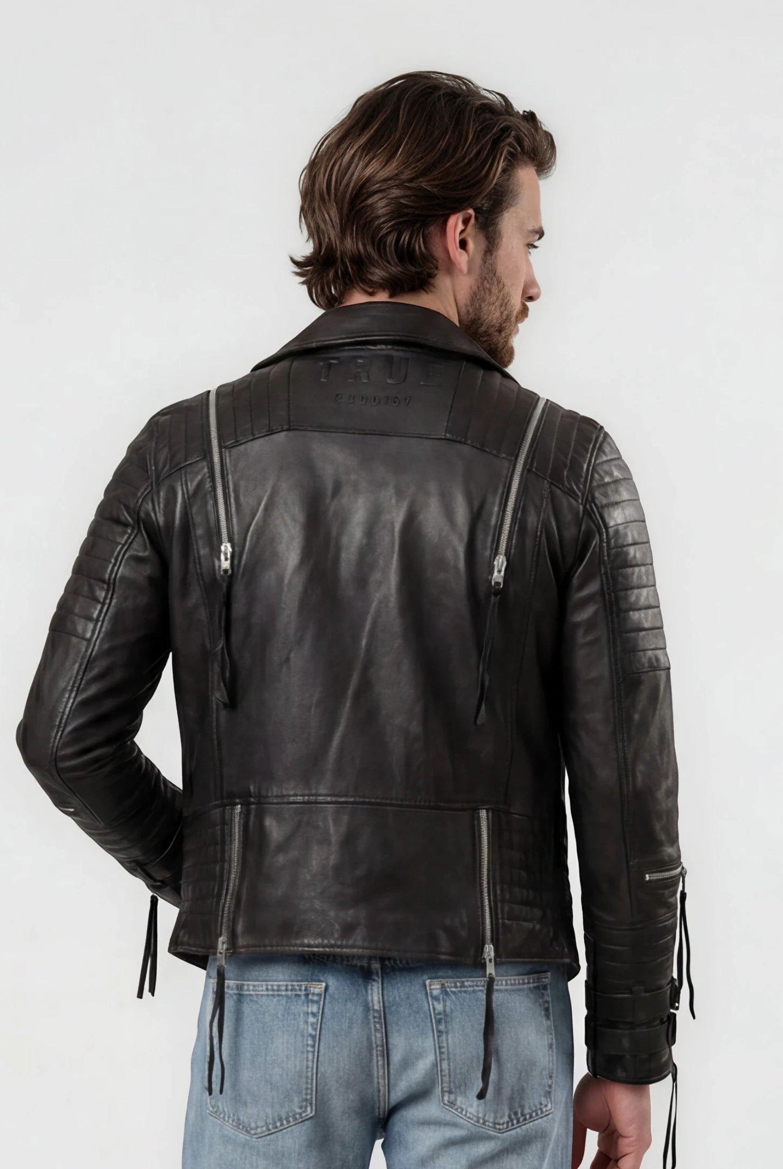 Ein Mann mit braunem Haar steht mit dem Rücken zur Kamera und trägt die trueprodigy Biker Lederjacke Herren "Jackson" mit Schnallen und hellblaue Jeans vor einem schlichten weißen Hintergrund.