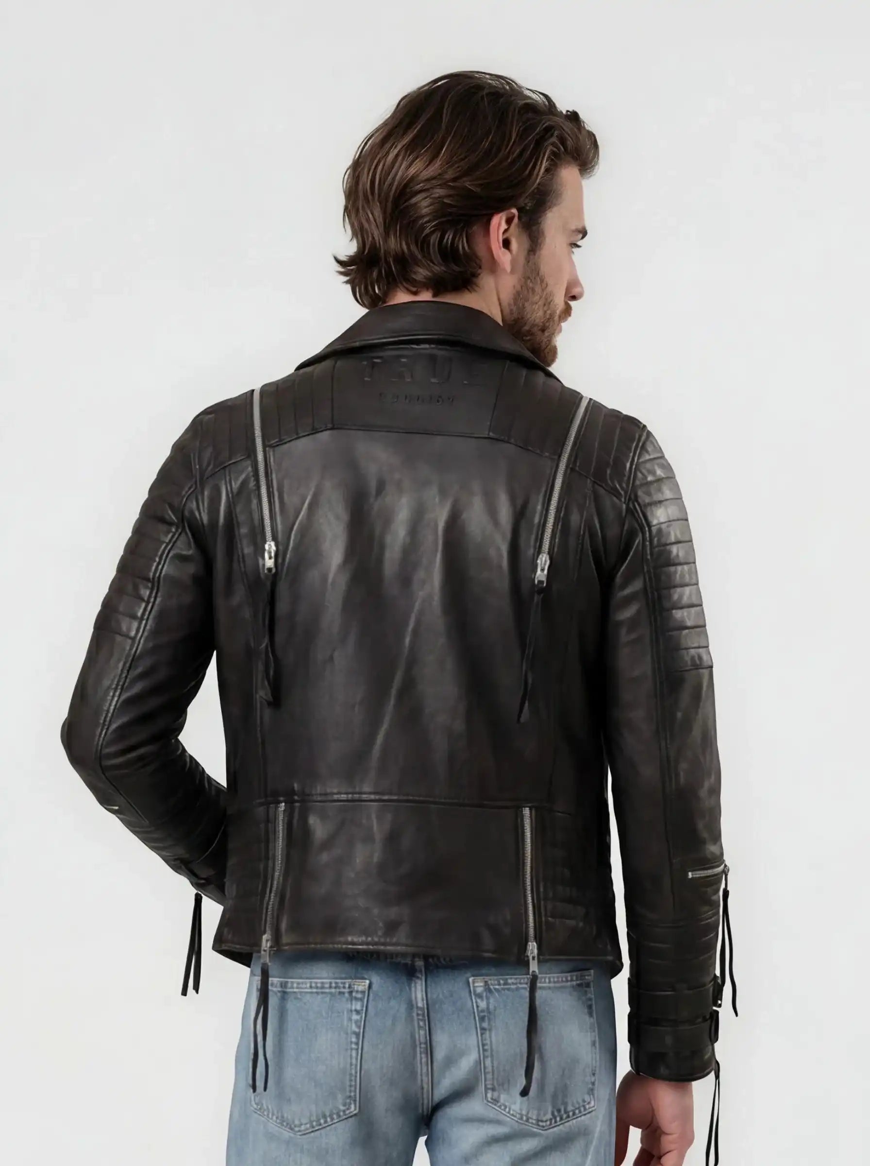 Ein Mann mit braunem Haar steht mit dem Rücken zur Kamera und trägt die trueprodigy Biker Lederjacke Herren "Jackson" mit Schnallen und hellblaue Jeans vor einem schlichten weißen Hintergrund.