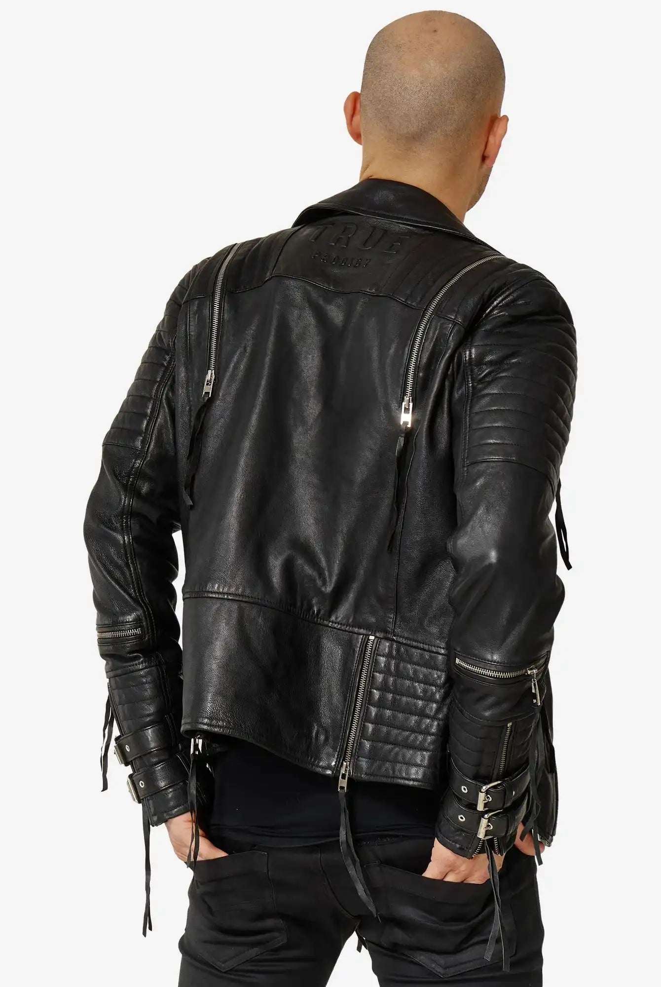 Eine Person mit rasiertem Kopf wird von hinten in einer schwarzen Jackson Bikerlederjacke mit Schnallen von trueprodigy, gepaart mit einer schwarzen Hose, vor einem schlichten weißen Hintergrund gezeigt.