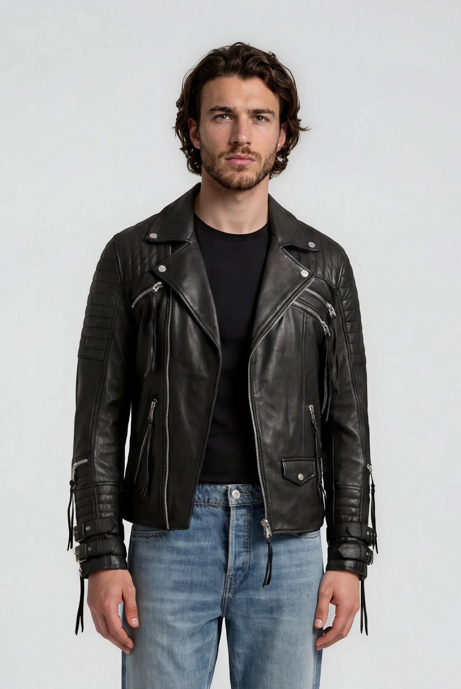 Ein Mann mit gewellten braunen Haaren und einem Bart trägt die trueprodigy Biker Lederjacke Herren "Jackson" mit Schnallen über einem schwarzen T-Shirt und hellblauen Jeans und steht vor einem schlichten hellgrauen Hintergrund mit Blick in die Kamera.