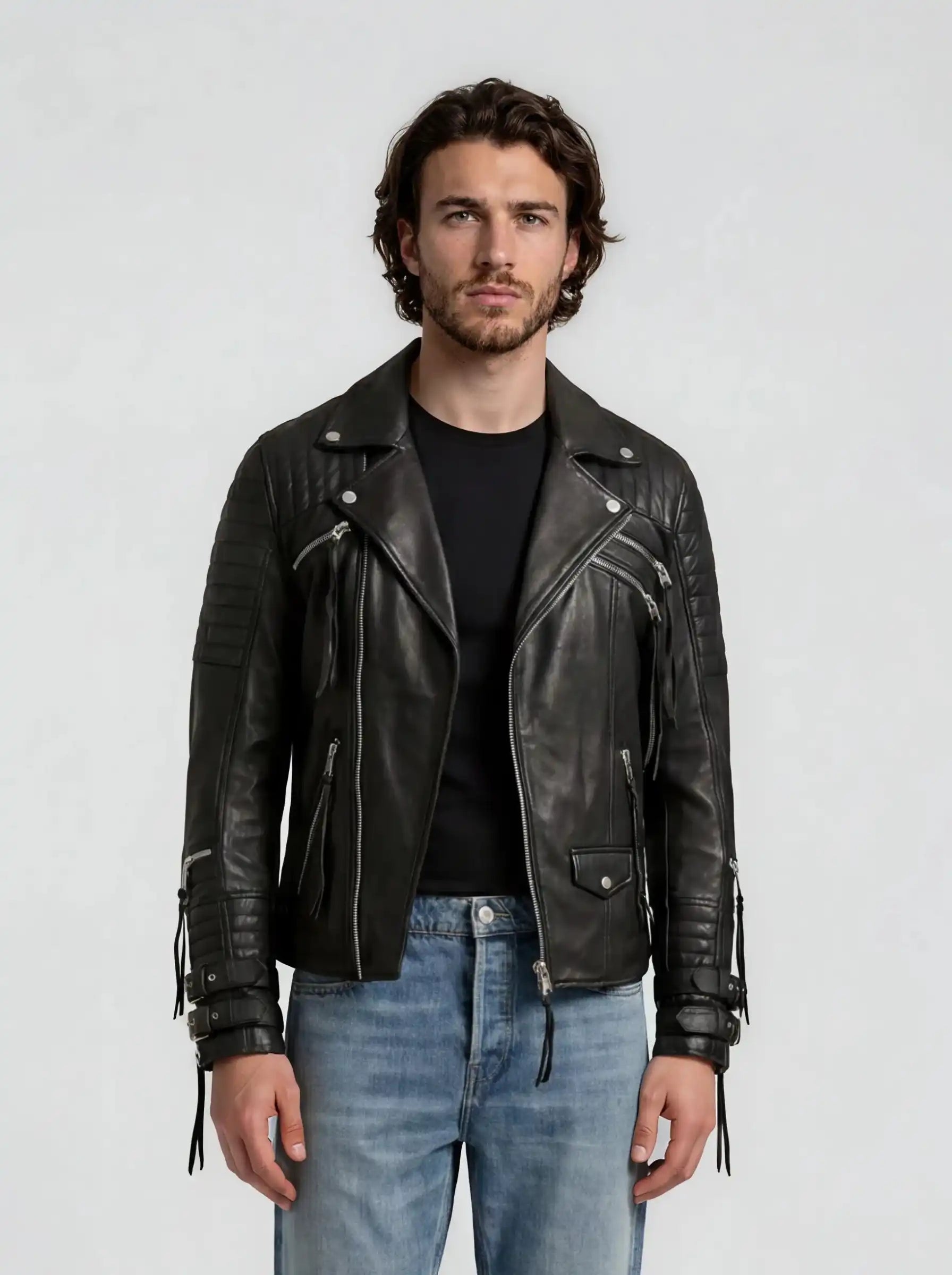 Ein Mann mit gewellten braunen Haaren und einem Bart trägt die trueprodigy Biker Lederjacke Herren "Jackson" mit Schnallen über einem schwarzen T-Shirt und hellblauen Jeans und steht vor einem schlichten hellgrauen Hintergrund mit Blick in die Kamera.
