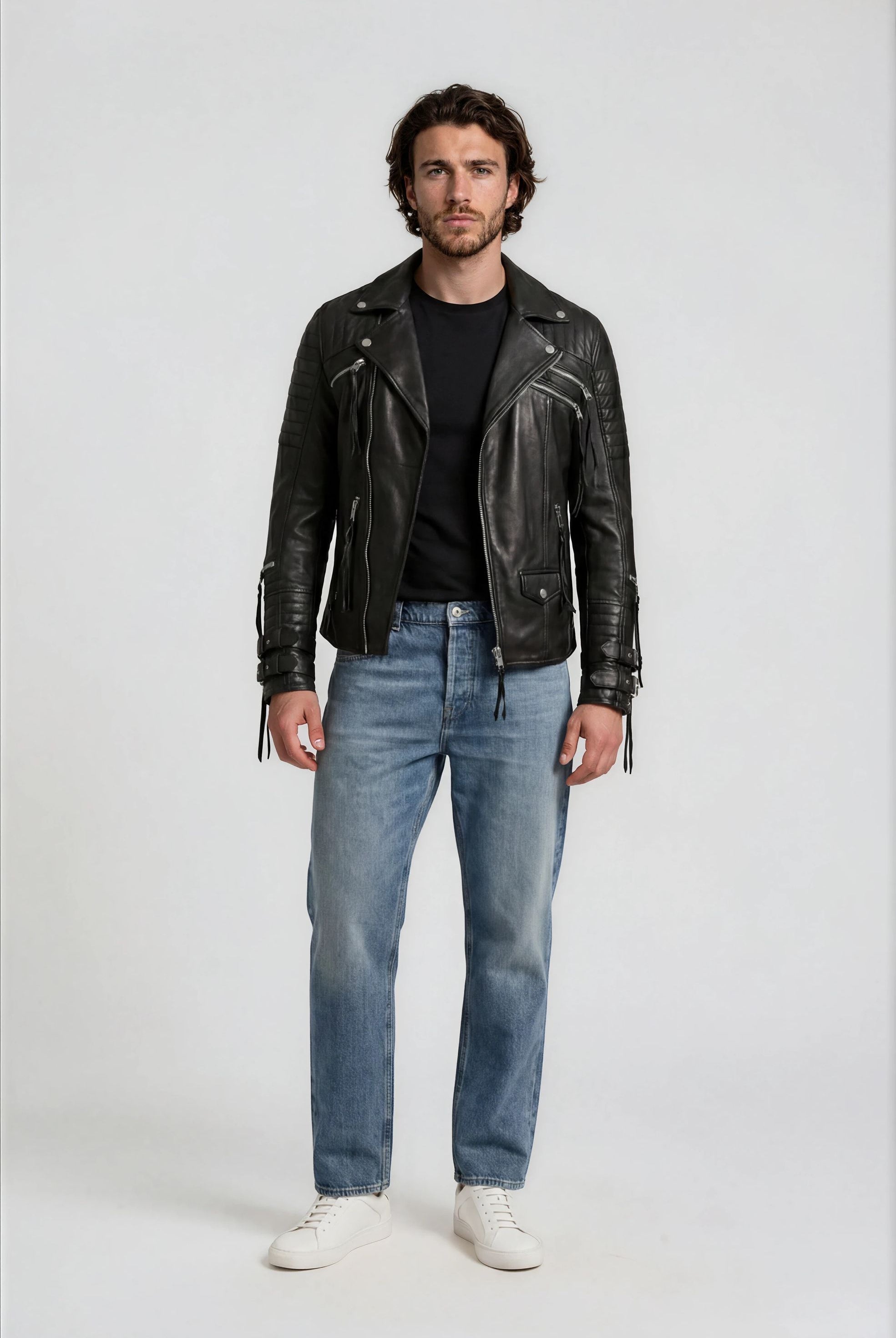 Ein Mann mit mittellangem Haar und Bart steht mit dem Gesicht nach vorne in der trueprodigy Biker Lederjacke Herren "Jackson" mit Schnallen, gepaart mit einem schwarzen T-Shirt, hellblauen Jeans und weißen Turnschuhen vor einem schlichten hellen Hintergrund.