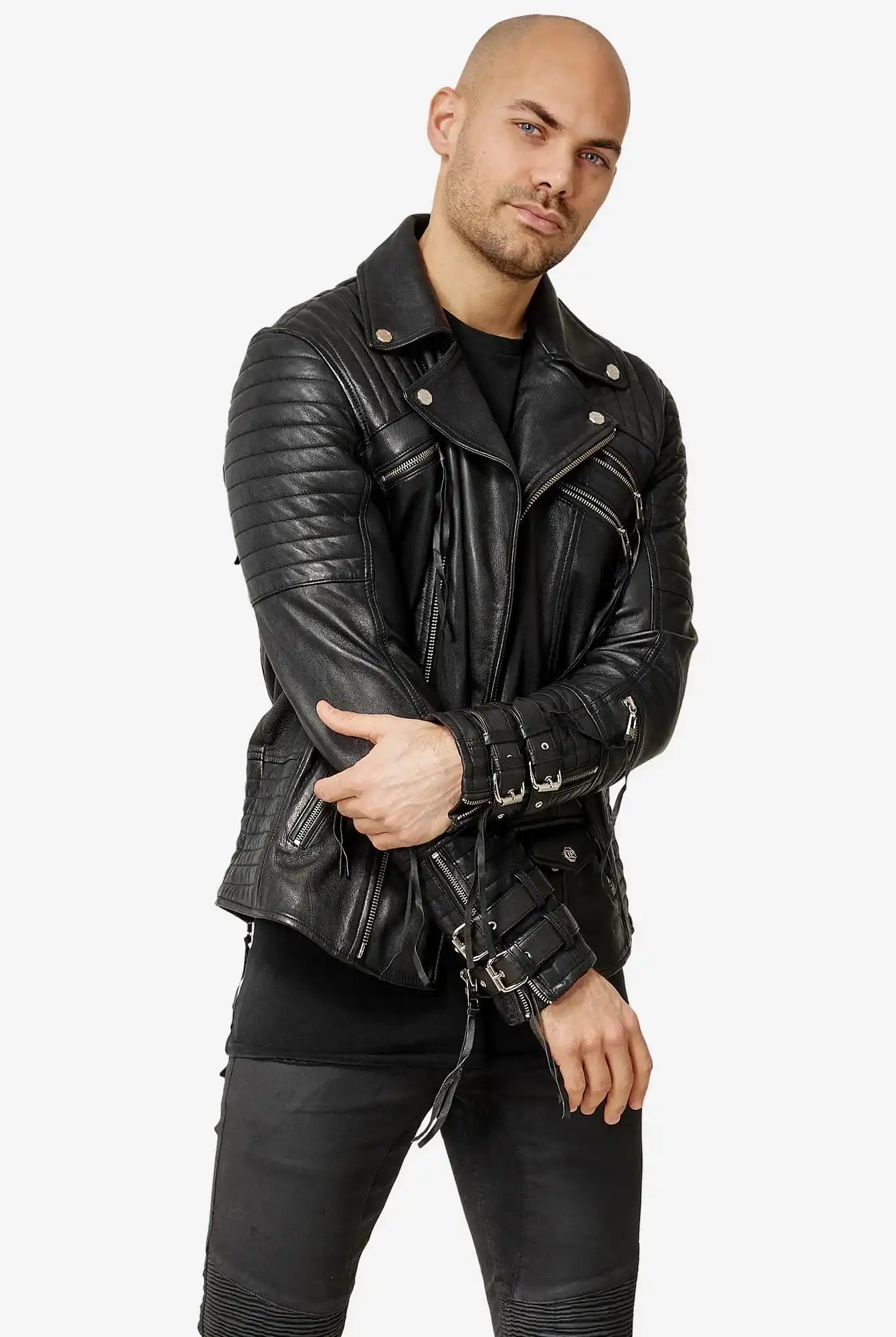 Eine Person mit rasiertem Kopf, bekleidet mit einer schwarzen Hose und der Trueprodigy Jackson Bikerlederjacke mit Schnallen, rückt den Ärmel der Jacke zurecht und blickt dabei in die Kamera vor einem schlichten weißen Hintergrund.