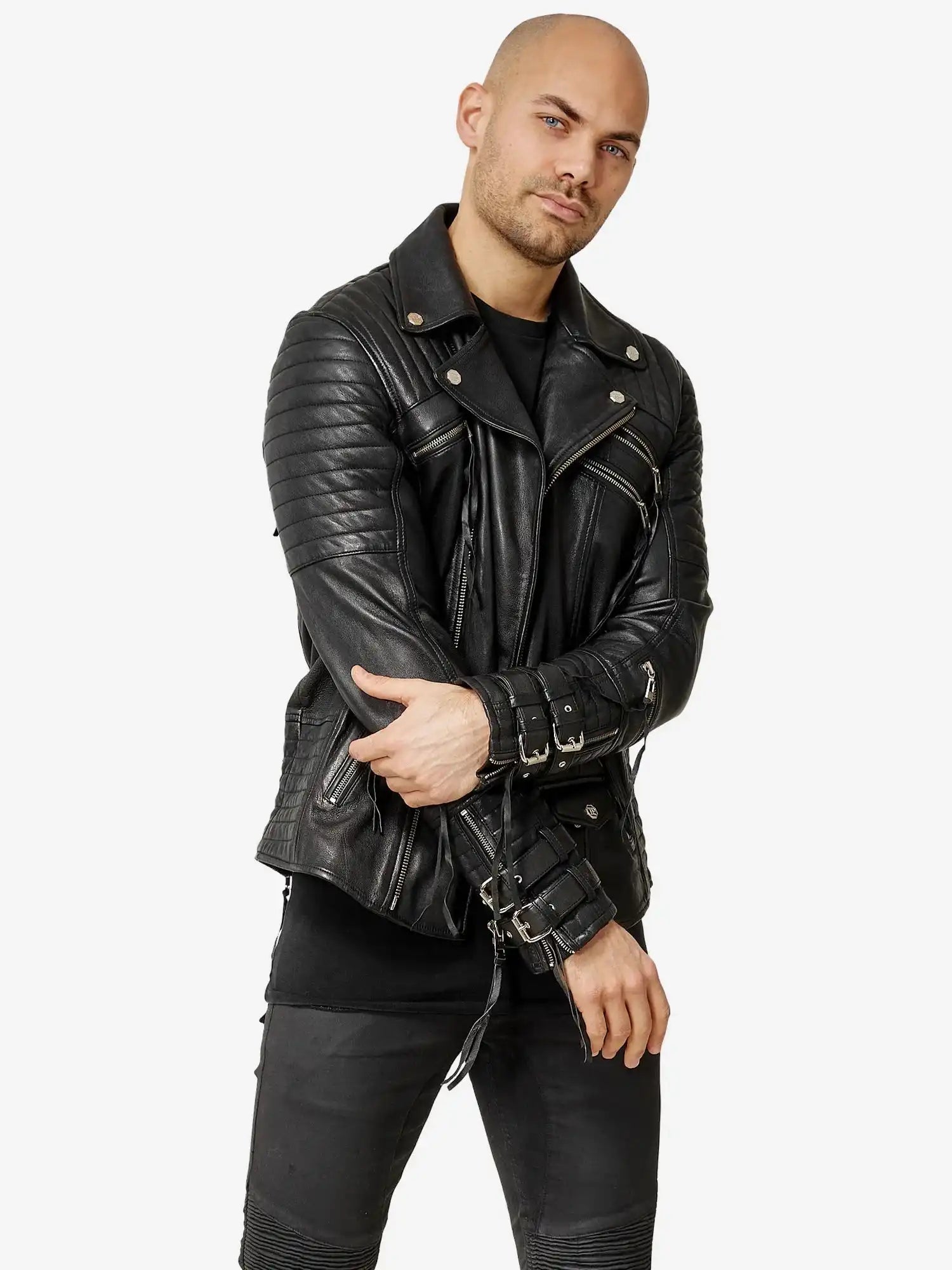 Eine Person mit rasiertem Kopf, bekleidet mit einer schwarzen Hose und der Trueprodigy Jackson Bikerlederjacke mit Schnallen, rückt den Ärmel der Jacke zurecht und blickt dabei in die Kamera vor einem schlichten weißen Hintergrund.
