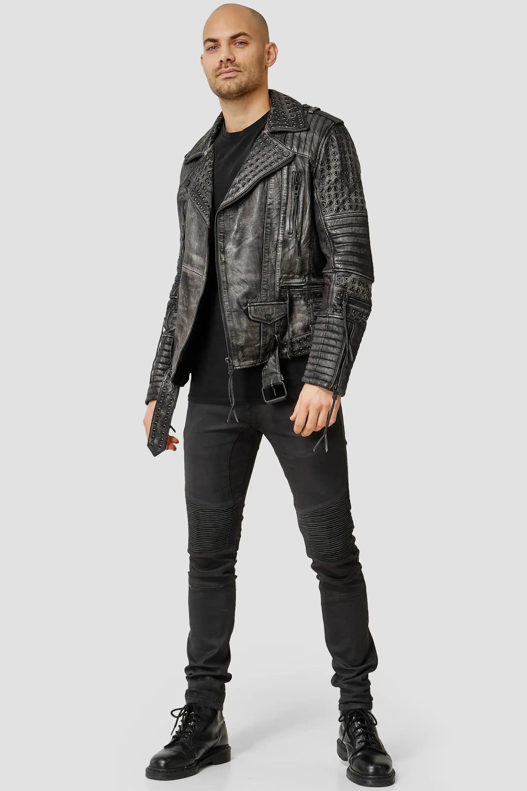 Ein Mann blickt nach vorne und trägt die Geralt Vintage Lederjacke mit Ösen von trueprodigy mit schwarzem Finish im Used-Look und strukturierten Details, gepaart mit einem schwarzen T-Shirt, einer schwarzen Hose und Schnürstiefeln vor einem hellgrauen Hintergrund.