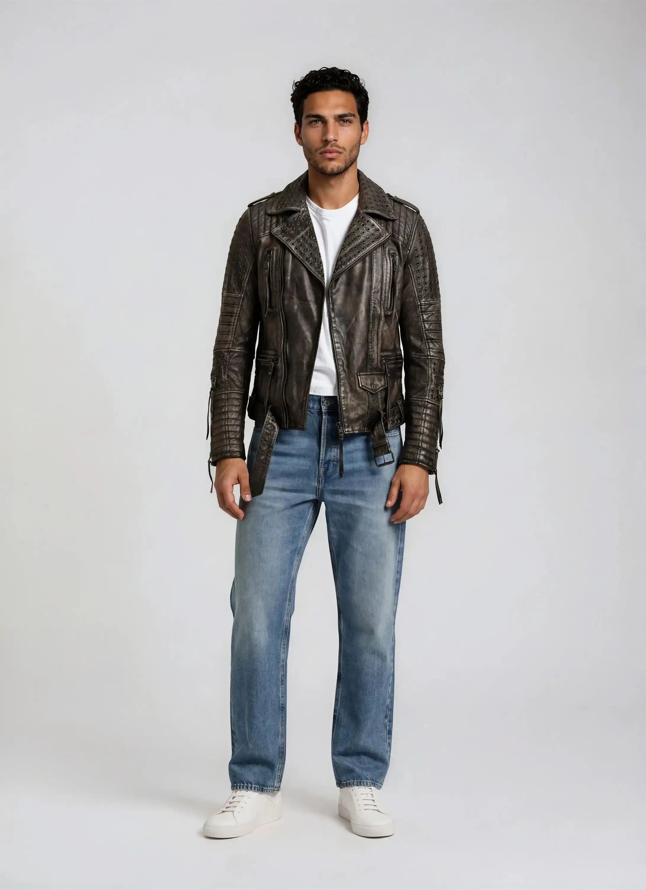 Ein Mann steht vor einem schlichten, hellen Hintergrund. Er trägt die trueprodigy Vintage Biker Lederjacke Herren "Geralt" mit Ösen in distressed brown, ein weißes T-Shirt, Blue Jeans und weiße Turnschuhe.