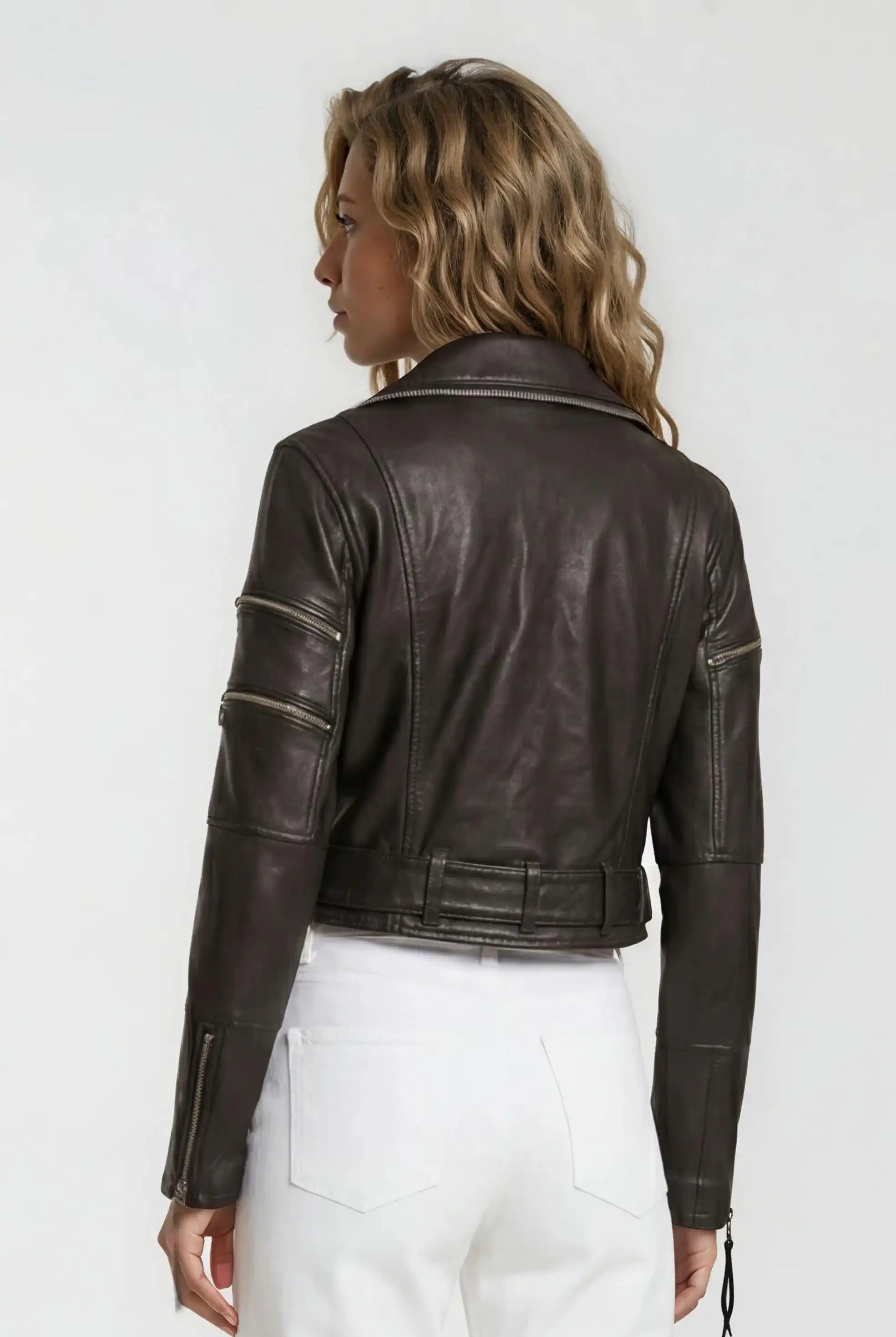 Eine Person mit gewelltem blondem Haar ist von hinten zu sehen. Sie trägt eine Trueprodigy Bikerlederjacke mit Gürtel mit Reißverschluss und eine weiße Hose vor einem schlichten hellen Hintergrund.