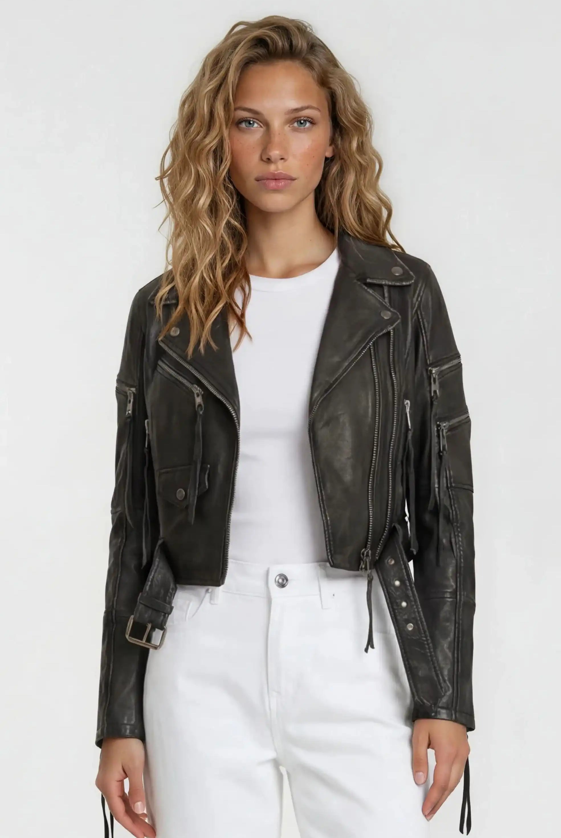 Eine Frau mit welligem blondem Haar trägt die Bikerlederjacke mit Gürtel von trueprodigy über einem weißen T-Shirt und hoch taillierten weißen Hosen vor schlicht hellem Hintergrund.