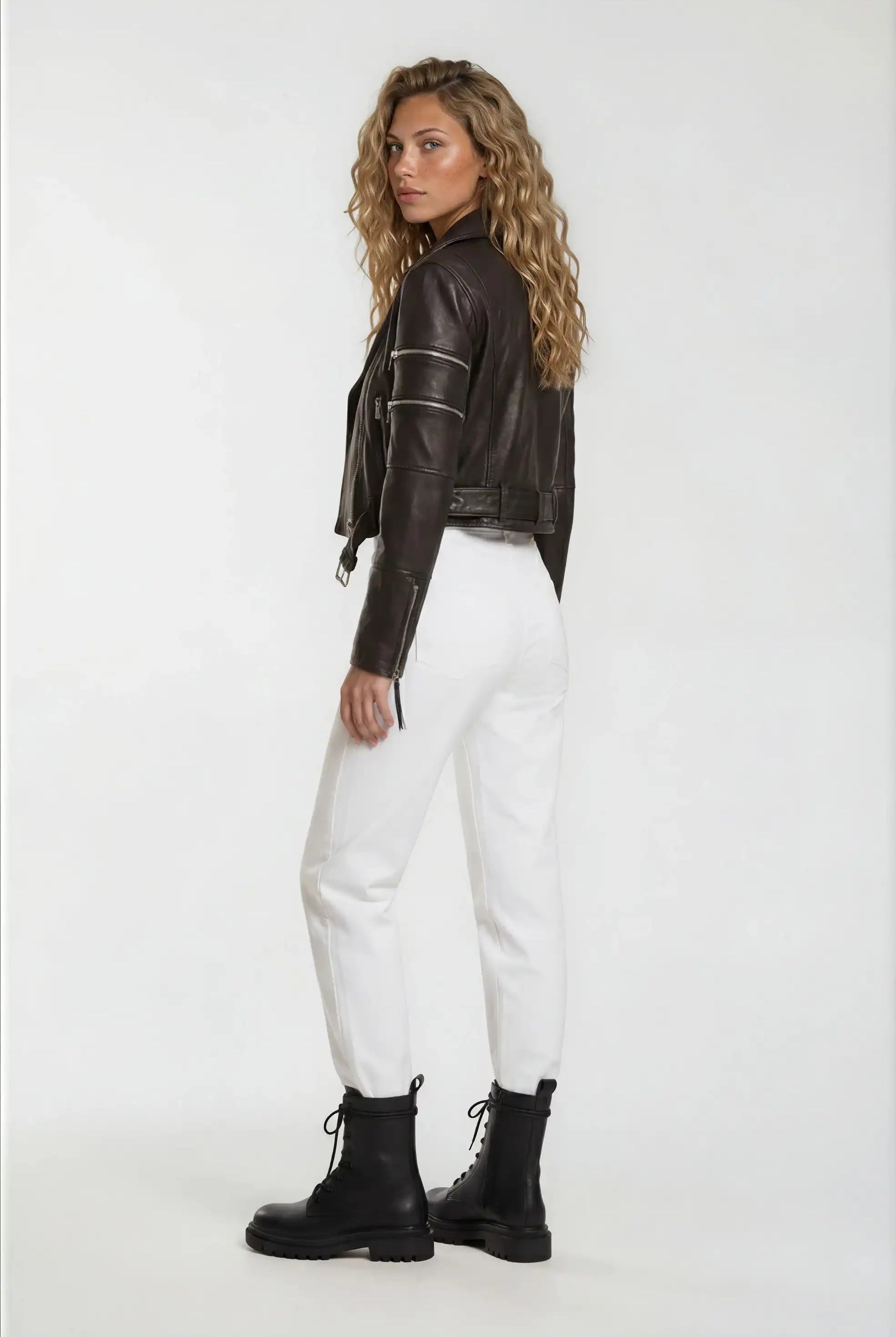 Eine Frau mit gewelltem blondem Haar steht leicht gedreht und trägt die trueprodigy Bikerlederjacke mit Gürtel aus Schafsleder, weißen Hosen und schwarzen Schnürstiefel vor einem schlichten weißen Hintergrund.