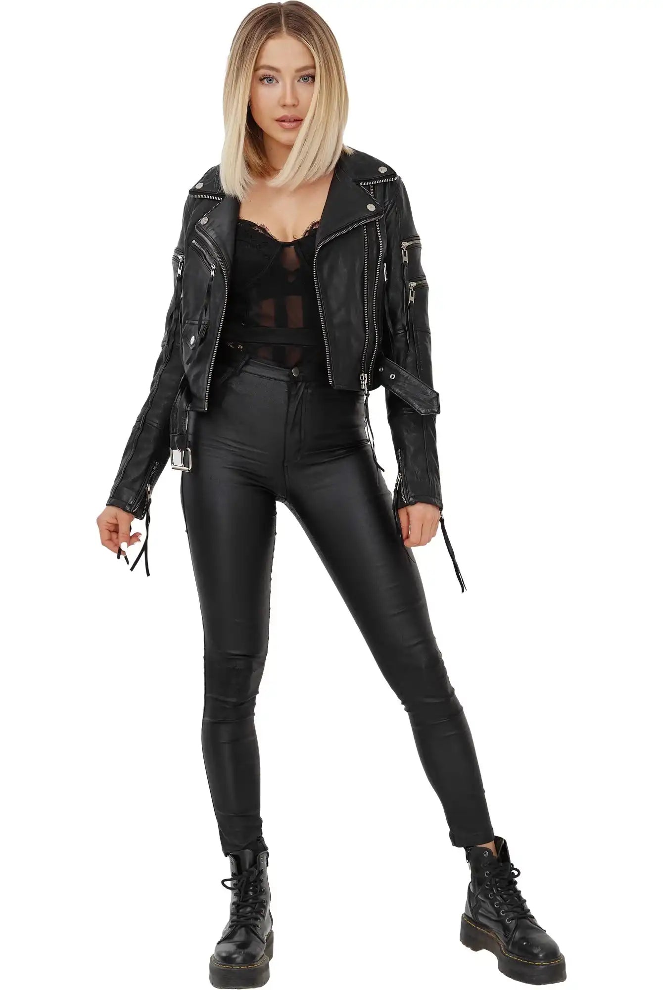 Eine Frau mit glattem, schulterlangem blondem Haar trägt die trueprodigy Izzy Bikerlederjacke mit Gürtel, ein schwarzes durchsichtiges Oberteil, eine hochtaillierte Hose und Kampfstiefel und posiert vor einem schlichten weißen Hintergrund.