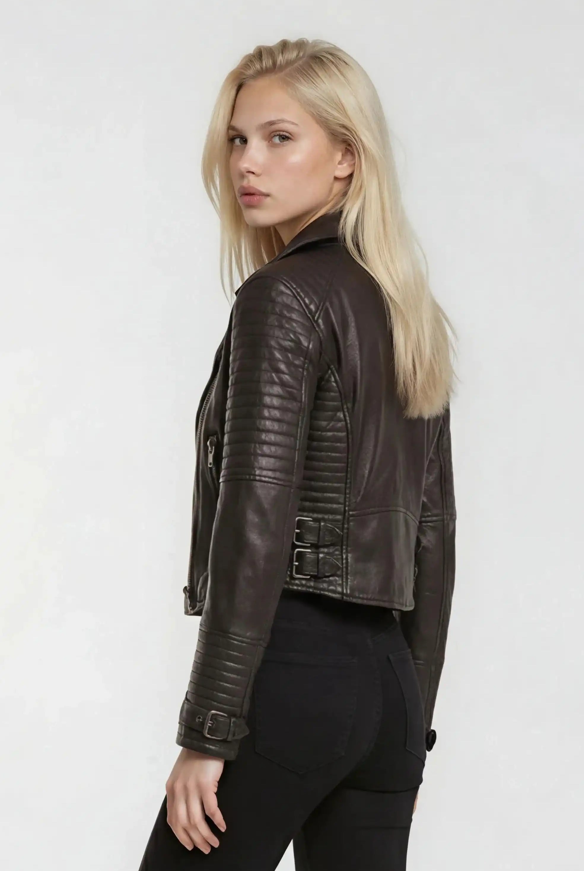 Eine Frau mit langen blonden Haaren steht leicht zur Seite gedreht, trägt eine trueprodigy Bikerlederjacke mit Schnallen und eine schwarze Hose und blickt über ihre Schulter in die Kamera vor einem schlichten hellen Hintergrund.