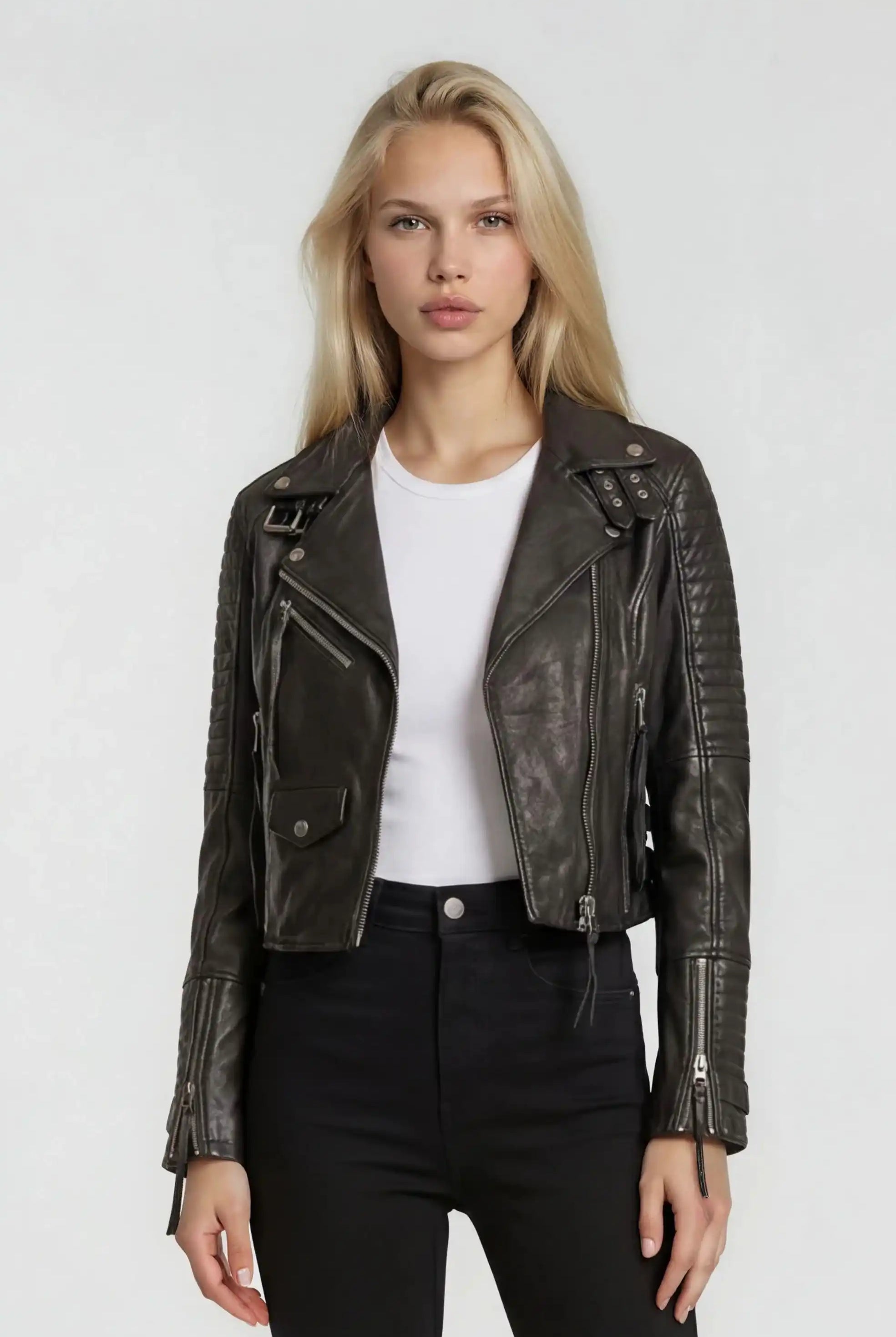 Eine Person mit langen blonden Haaren trägt die trueprodigy Bikerlederjacke mit Schnallen im klassischen Biker-Look über einem weißen T-Shirt und einer hochgezogenen schwarzen Hose, stehend vor einem schlichten hellen Hintergrund.
