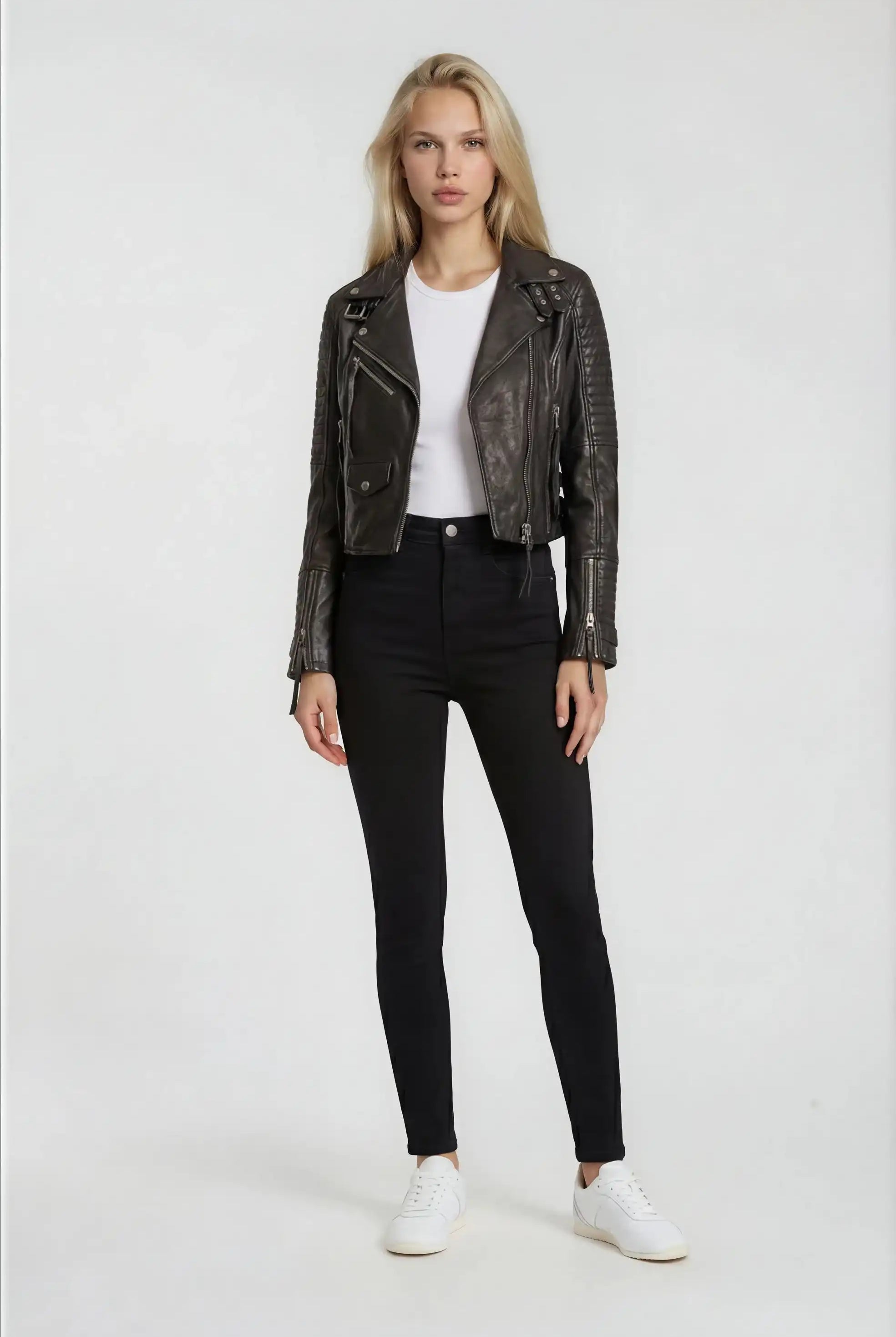 Eine Person mit langen blonden Haaren steht vor einem schlichten Hintergrund und trägt die Bikerlederjacke mit Schnallen von trueprodigy, gepaart mit einem weißen T-Shirt, einer schwarzen High-Waist-Hose und weißen Sneakers.