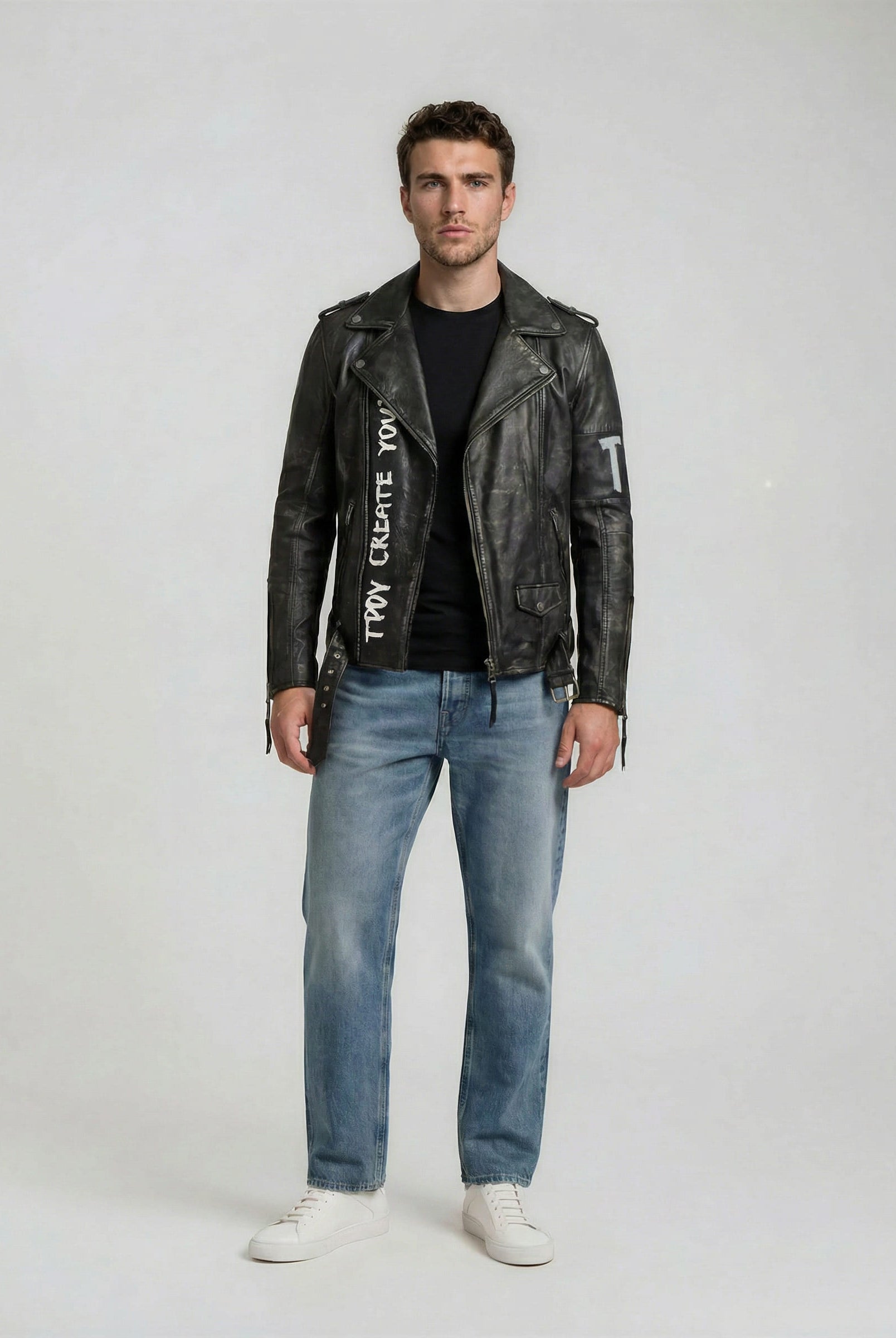 In der trueprodigy Biker Lederjacke Herren "Russell" mit Rücken-Print, gepaart mit einem schwarzen T-Shirt, hellblauen Jeans und weißen Turnschuhen, blickt ein Mann nach vorne und sorgt vor einem schlichten Hintergrund für einen coolen Biker-Look.