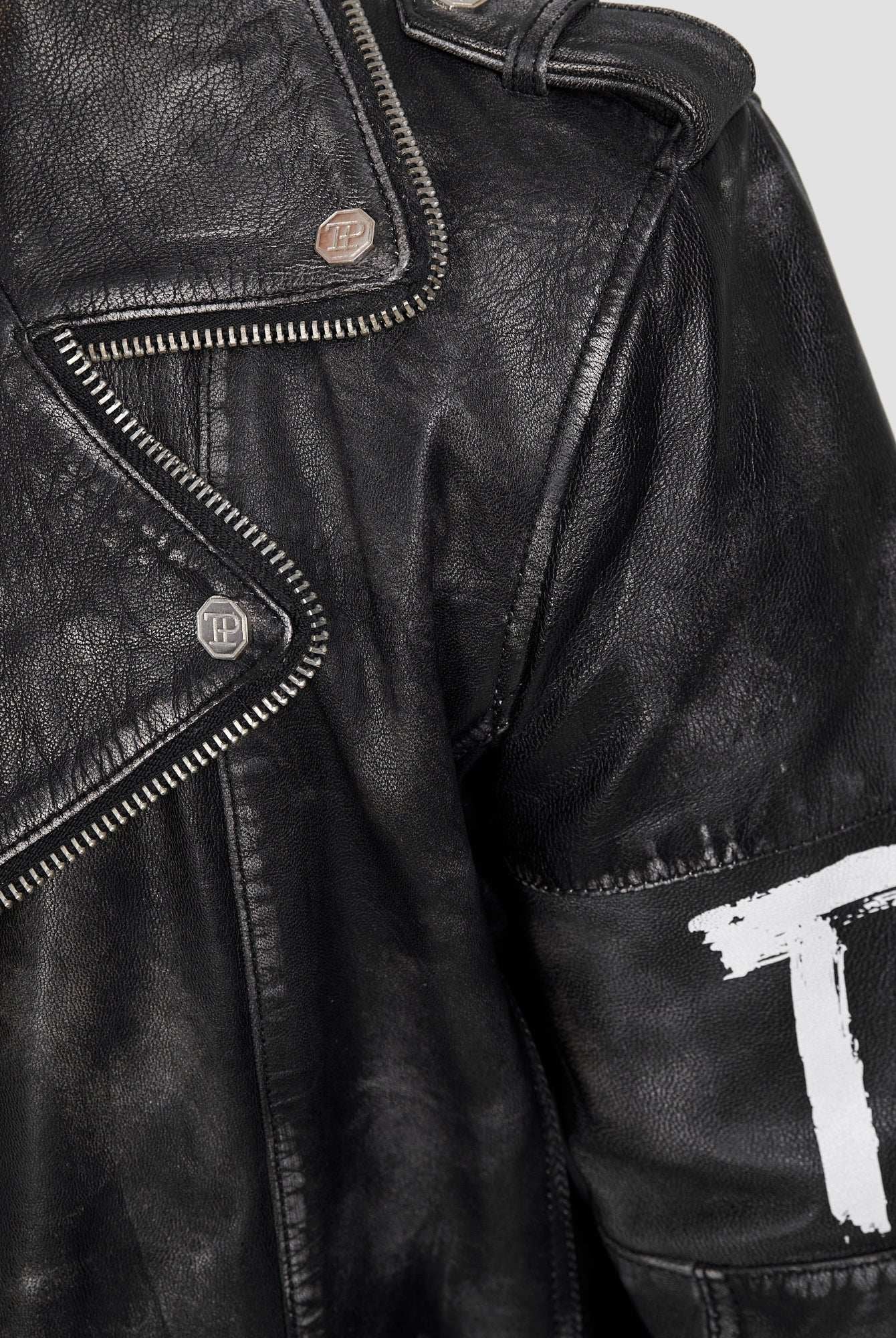 Nahaufnahme der trueprodigy Russell Lederjacke mit Print: schwarzes Ziegenleder, Metalldruckknöpfe, freiliegender silberner Reißverschluss, weiß lackiertes T auf dem Ärmel, und ein strukturierter, leicht abgenutzter Biker-Look.