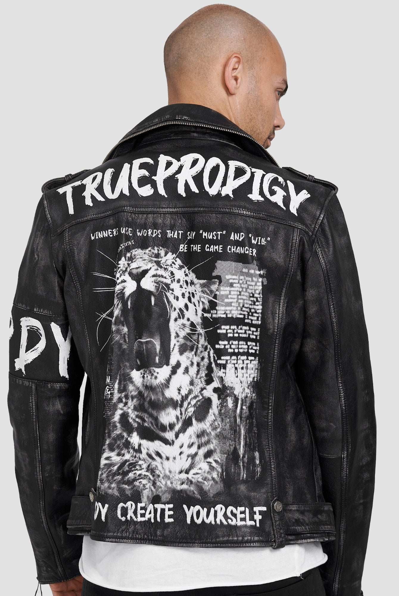 Ein glatzköpfiger Mann, der auf die Seite gedreht ist, trägt die trueprodigy Russell Lederjacke mit Print in klassischem Schwarz, mit fetter weißer Schrift und einer brüllenden Leopardengrafik auf dem Rücken, für einen wilden Biker-Look mit Slogans wie TRUE PRODIGY und CREATE YOURSELF.