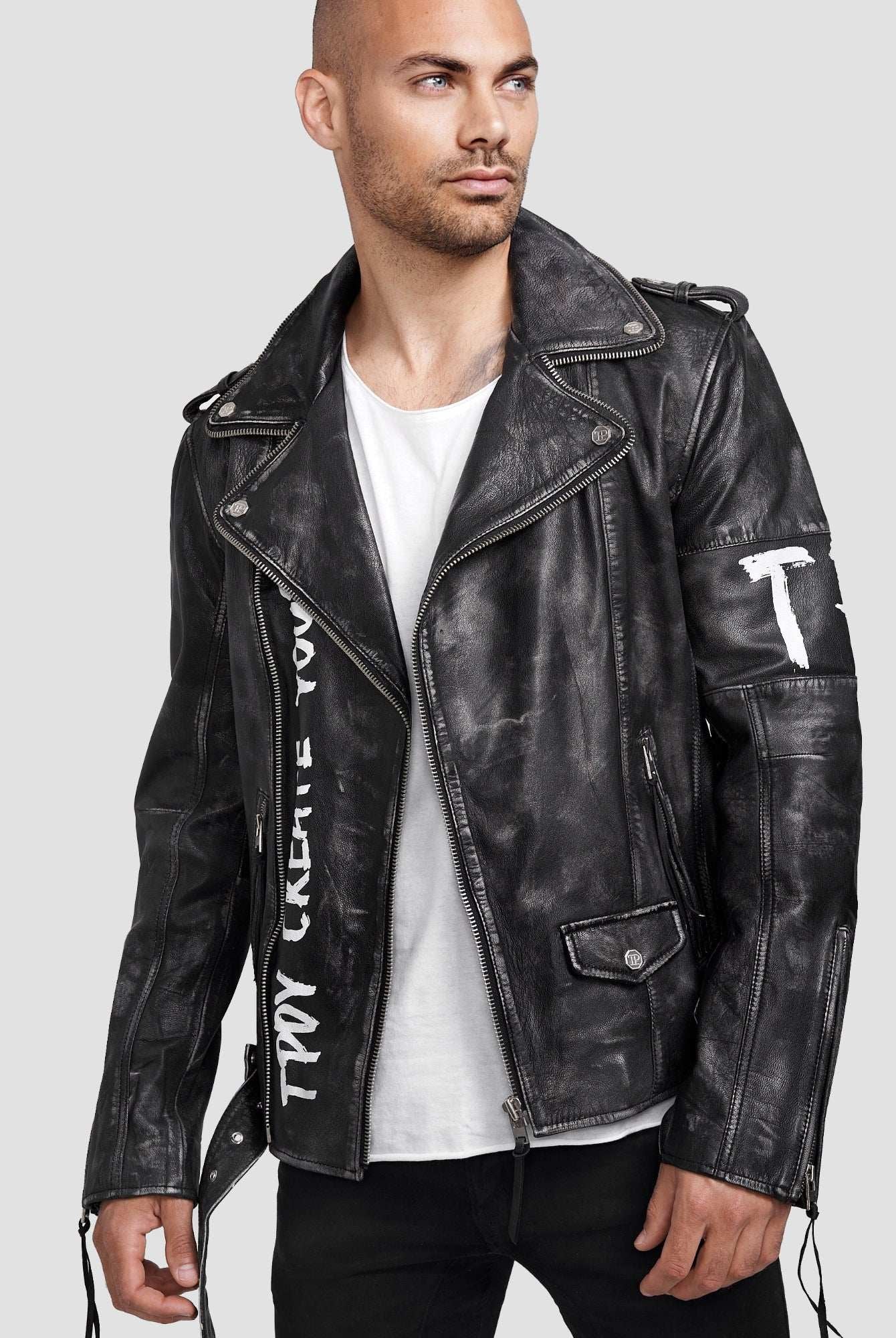 Ein Mann in der trueprodigy Russell Lederjacke mit Print - einer schwarzen Lederjacke im Biker-Stil mit weißem Graffiti-Schriftzug -, einem weißen T-Shirt und einer schwarzen Hose steht vor einem schlichten hellgrauen Hintergrund.