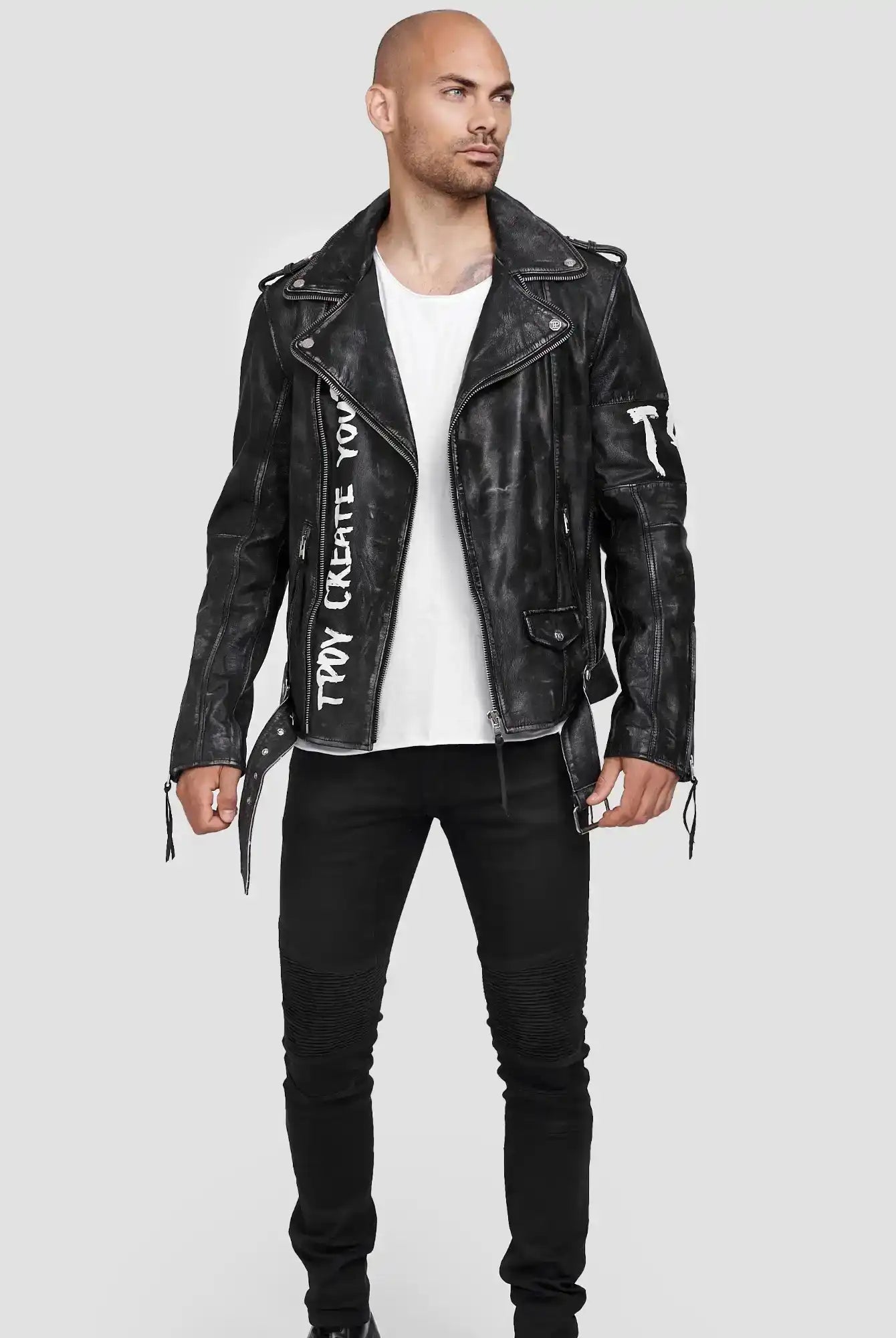 Ein Mann steht vor einem hellgrauen Hintergrund und trägt die Russell Lederjacke mit Print aus Ziegenleder von trueprodigy, gepaart mit einem weißen T-Shirt, schwarzen Jeans und schwarzen Schuhen. Seine Hände liegen entspannt an seinen Seiten, während er leicht nach links schaut.