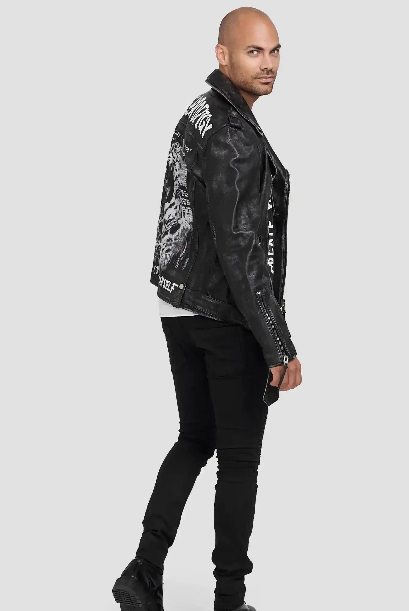 Ein Mann mit rasiertem Kopf trägt die schwarze Russell Lederjacke mit Print von trueprodigy, gepaart mit schwarzer Hose und Stiefeln im coolen Biker-Look, zur Seite gedreht und über die Schulter schauend vor einem hellgrauen Hintergrund.
