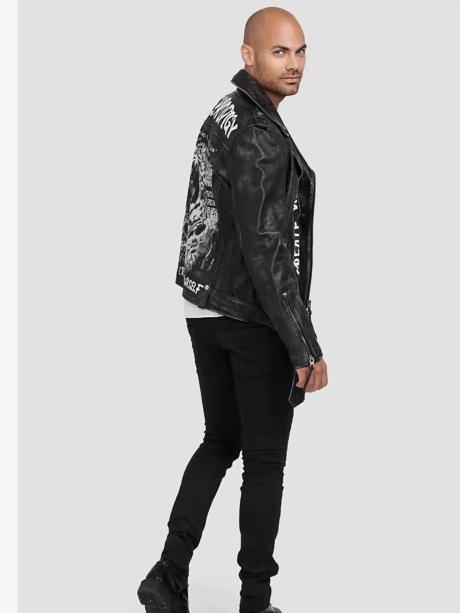Ein Mann mit rasiertem Kopf trägt die schwarze Russell Lederjacke mit Print von trueprodigy, gepaart mit schwarzer Hose und Stiefeln im coolen Biker-Look, zur Seite gedreht und über die Schulter schauend vor einem hellgrauen Hintergrund.