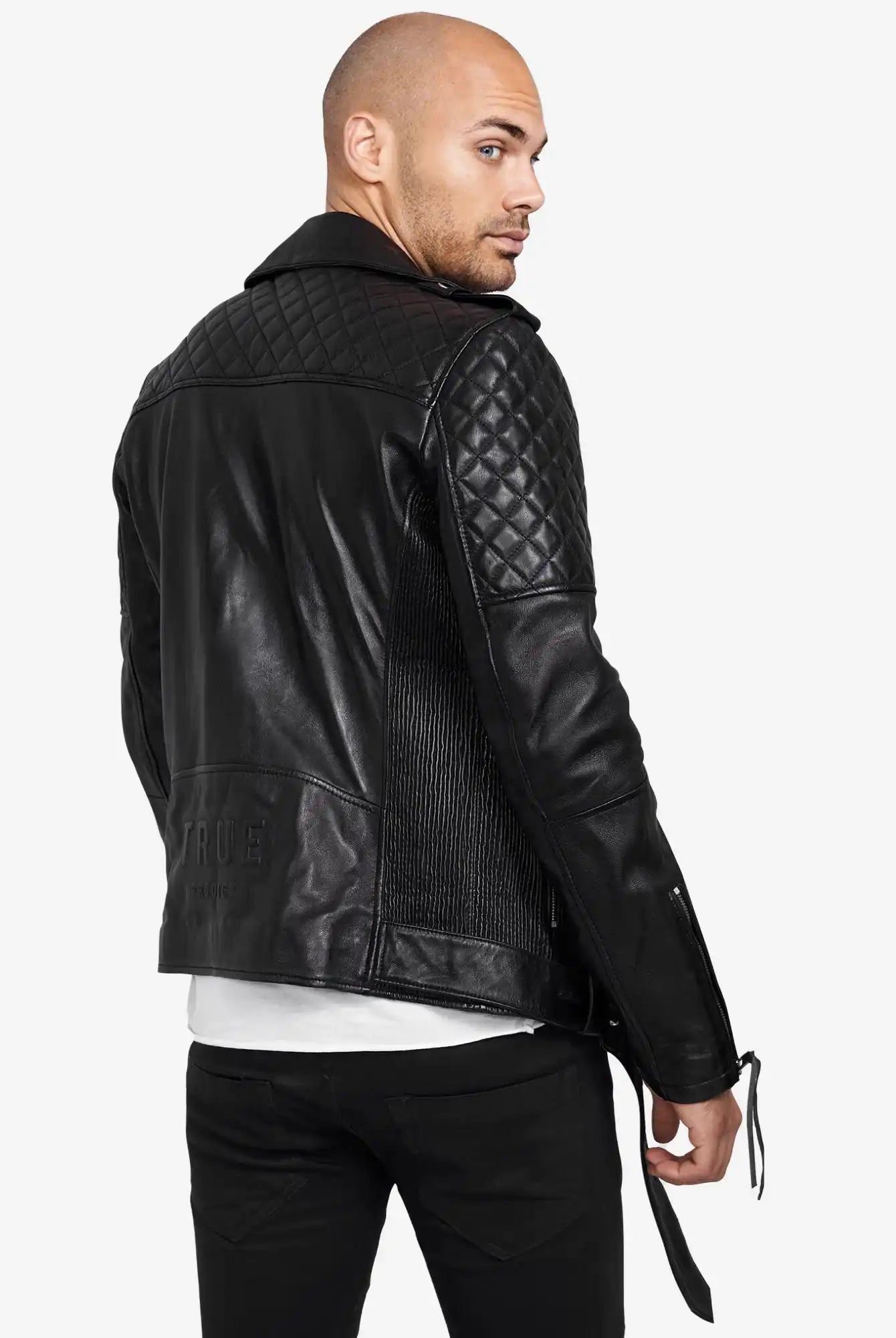 Ein Mann mit rasiertem Kopf modelliert die trueprodigy Simba Bikerlederjacke mit Gürtel in schwarzem Leder mit gesteppten Schultern und Ärmeln. Rückwärts gewandt blickt er über die linke Schulter und trägt eine schwarze Hose und ein weißes Hemd.