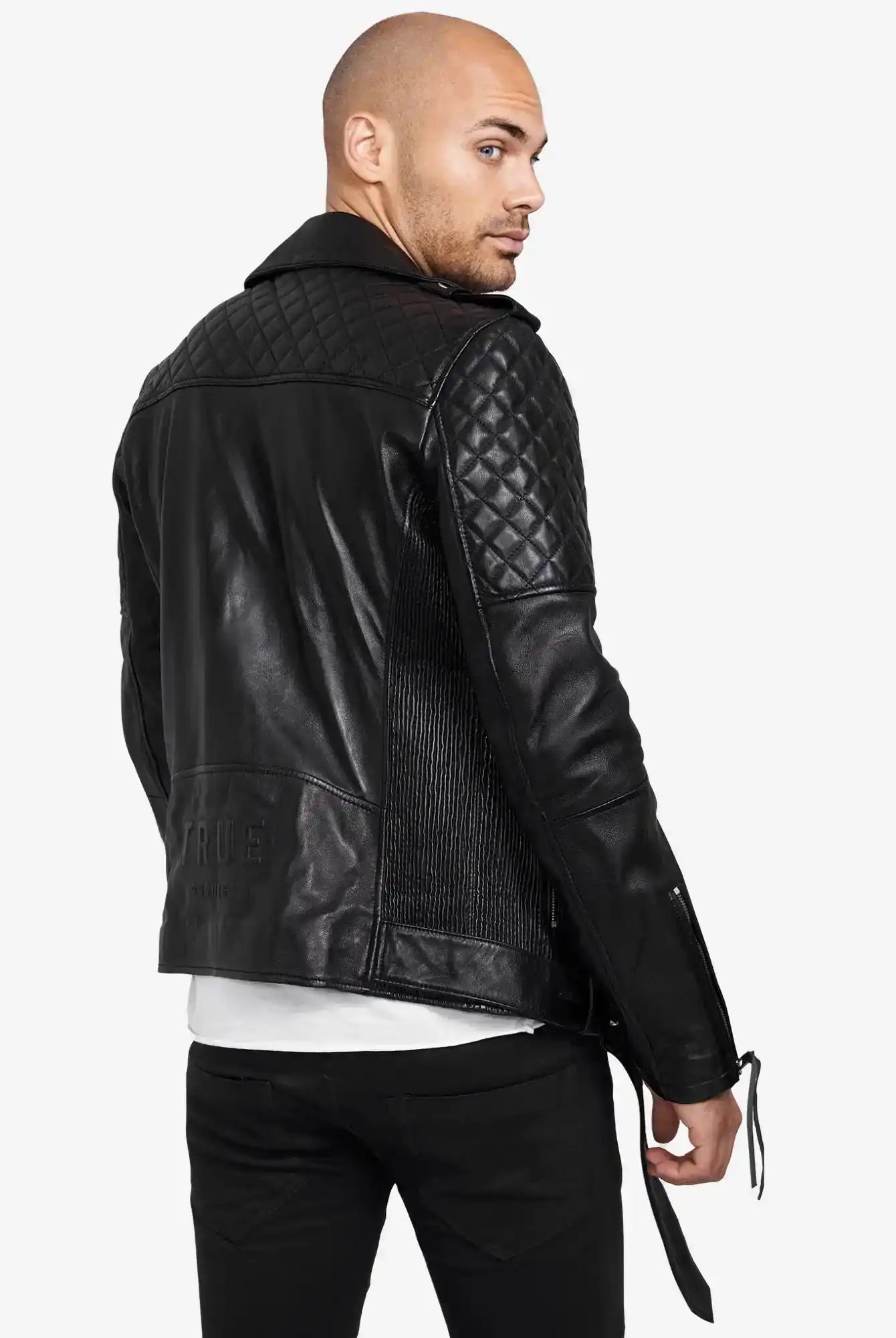 Ein Mann mit rasiertem Kopf modelliert die trueprodigy Simba Bikerlederjacke mit Gürtel in schwarzem Leder mit gesteppten Schultern und Ärmeln. Rückwärts gewandt blickt er über die linke Schulter und trägt eine schwarze Hose und ein weißes Hemd.